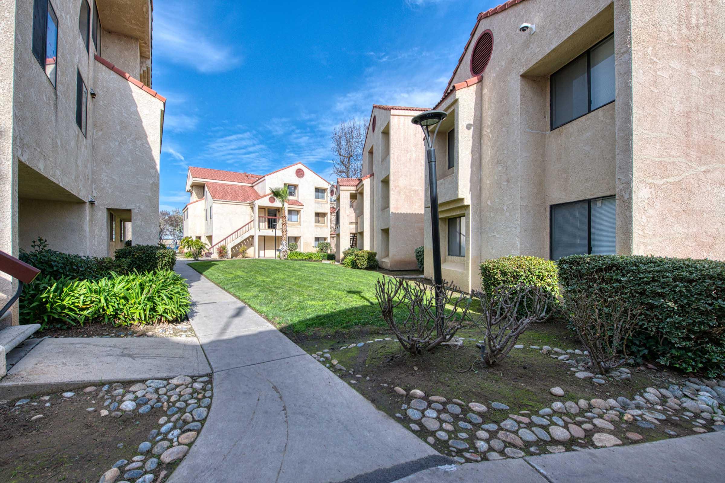 A view of an apartment complex with two-story buildings, a manicured lawn, and landscaped pathways. The blue sky is visible above, and the scene includes greenery and shrubs along the walkways, creating a welcoming residential atmosphere.