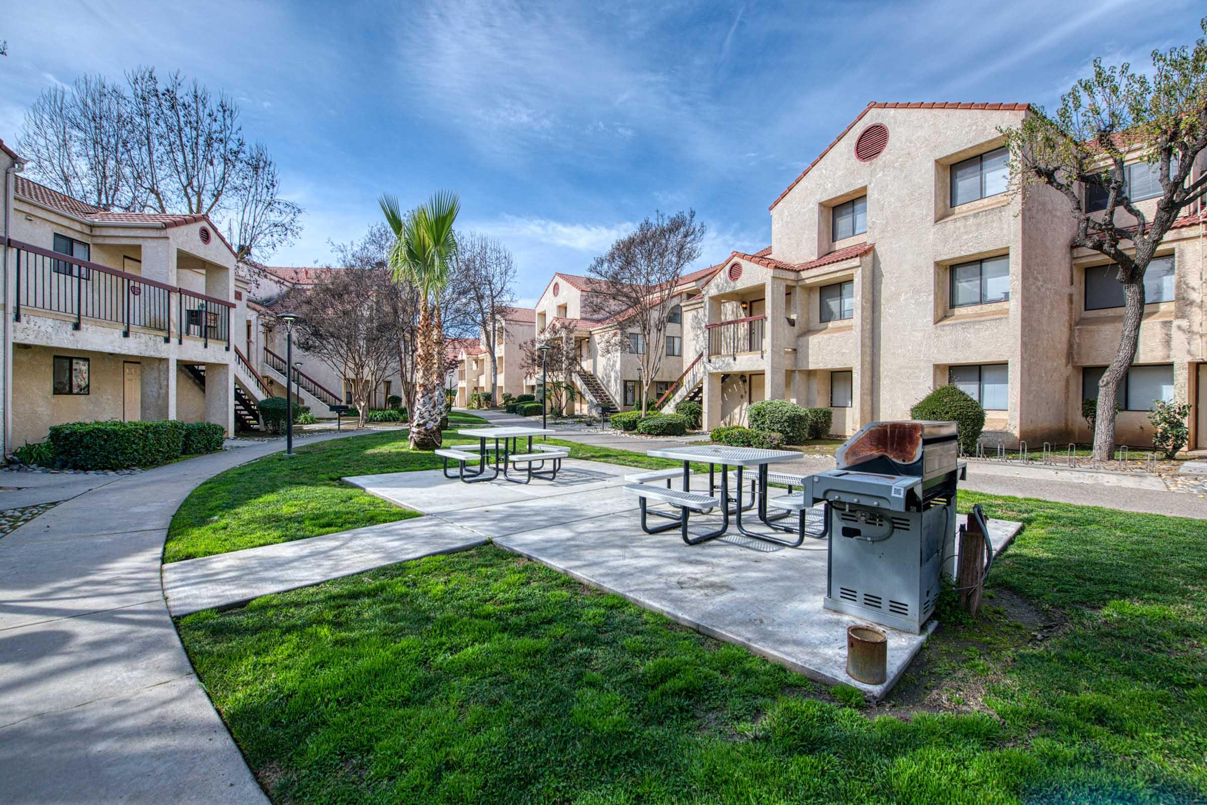 A well-maintained apartment complex courtyard featuring several buildings, green grass, picnic tables, a barbecue grill, and palm trees under a clear blue sky.