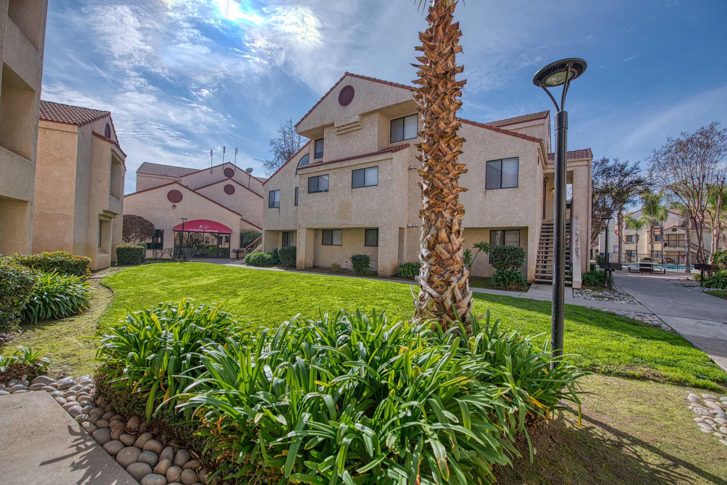 A well-kept residential complex featuring two-story buildings with a landscaped lawn, palm trees, and decorative rocks. The sky is clear with some clouds, and a lamp post is visible near the walkway. The scene captures a peaceful suburban atmosphere.