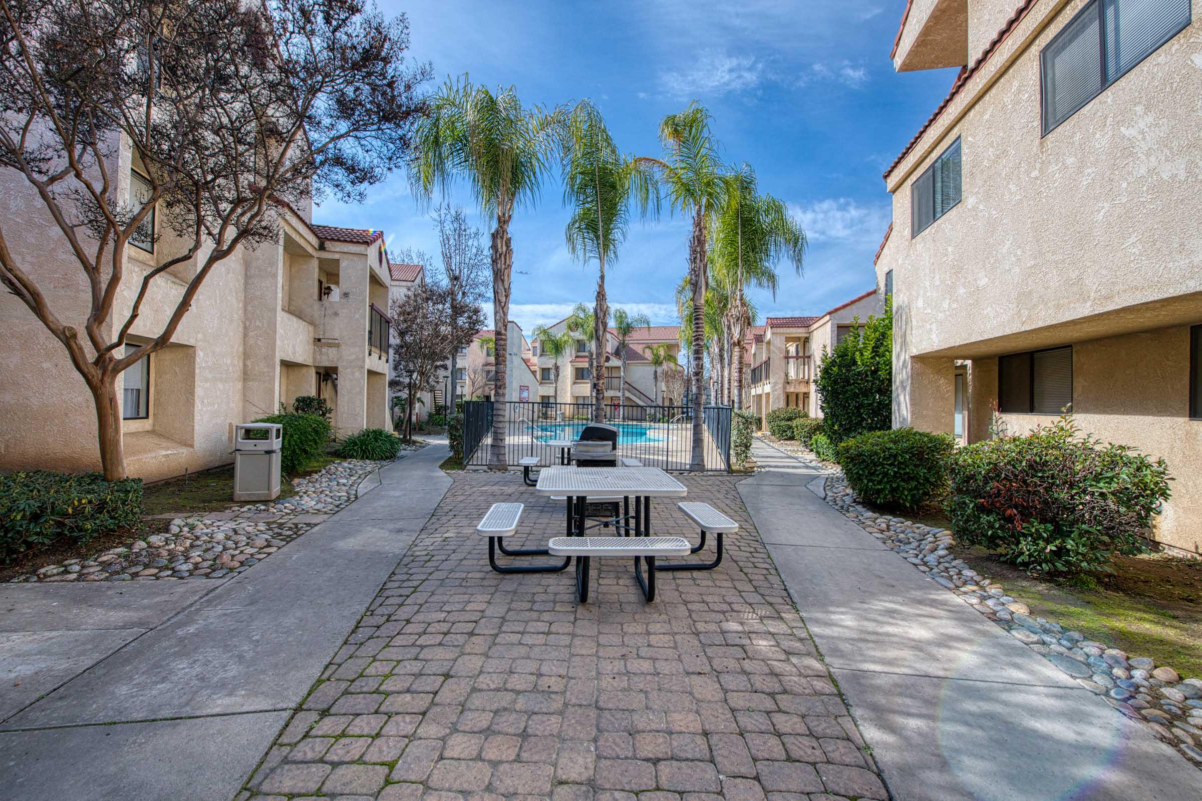 A courtyard area of an apartment complex featuring a picnic table, palm trees, and a swimming pool in the background. The pathways are paved with stones, and there are bushes and trees lining the sides of the buildings. The sky is clear and bright, creating a welcoming atmosphere.