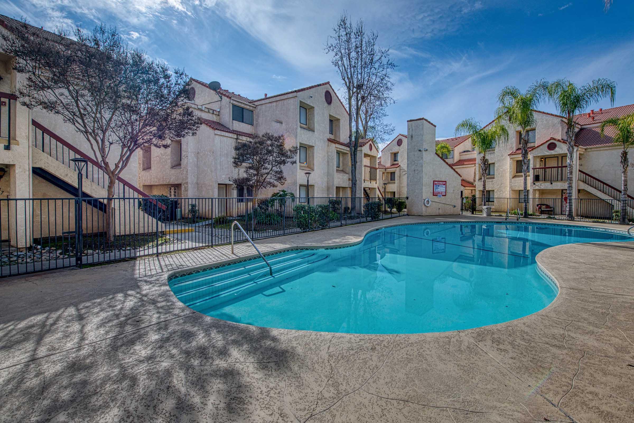 A sparkling blue swimming pool surrounded by a landscaped area, with two-story apartment buildings in the background. The scene features clear skies and lush greenery, creating a relaxing atmosphere for residents.
