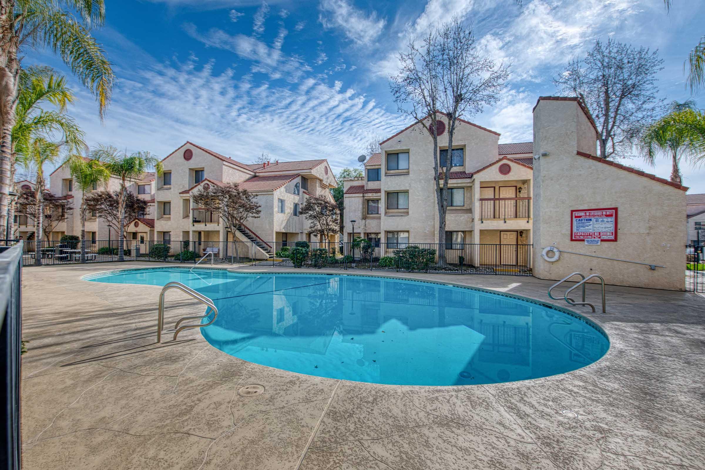 A clear swimming pool surrounded by a pool deck, with palm trees and landscaped areas. In the background, there are two-story apartment buildings with red accents, and a blue sky with wispy clouds overhead. A fence surrounds the pool area, enhancing the inviting atmosphere.