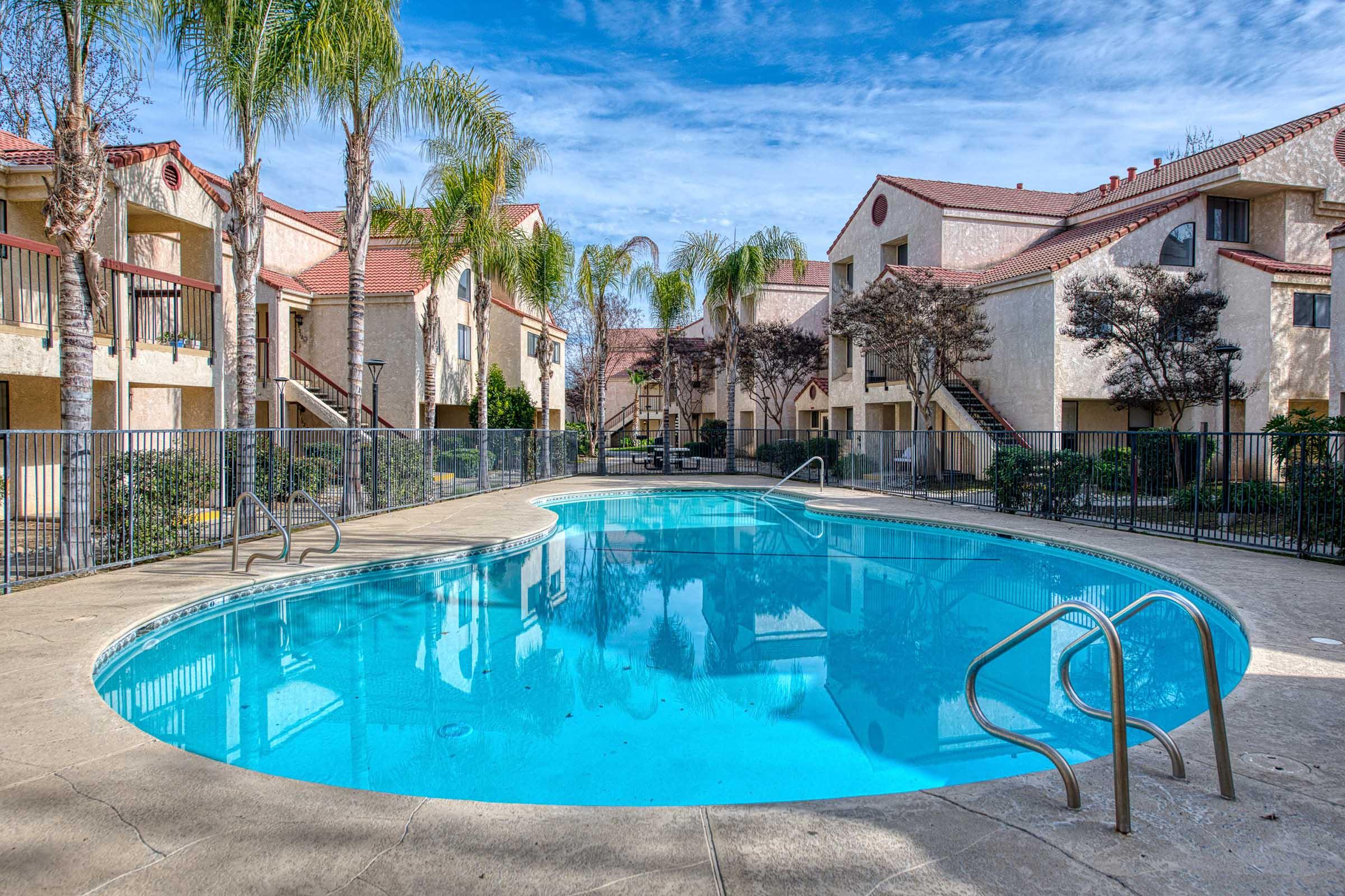 A clear blue swimming pool surrounded by palm trees and landscaped greenery, adjacent to two-story apartment buildings. The pool area features a smooth concrete deck and metal railings. Bright blue sky with scattered clouds completes the serene atmosphere.