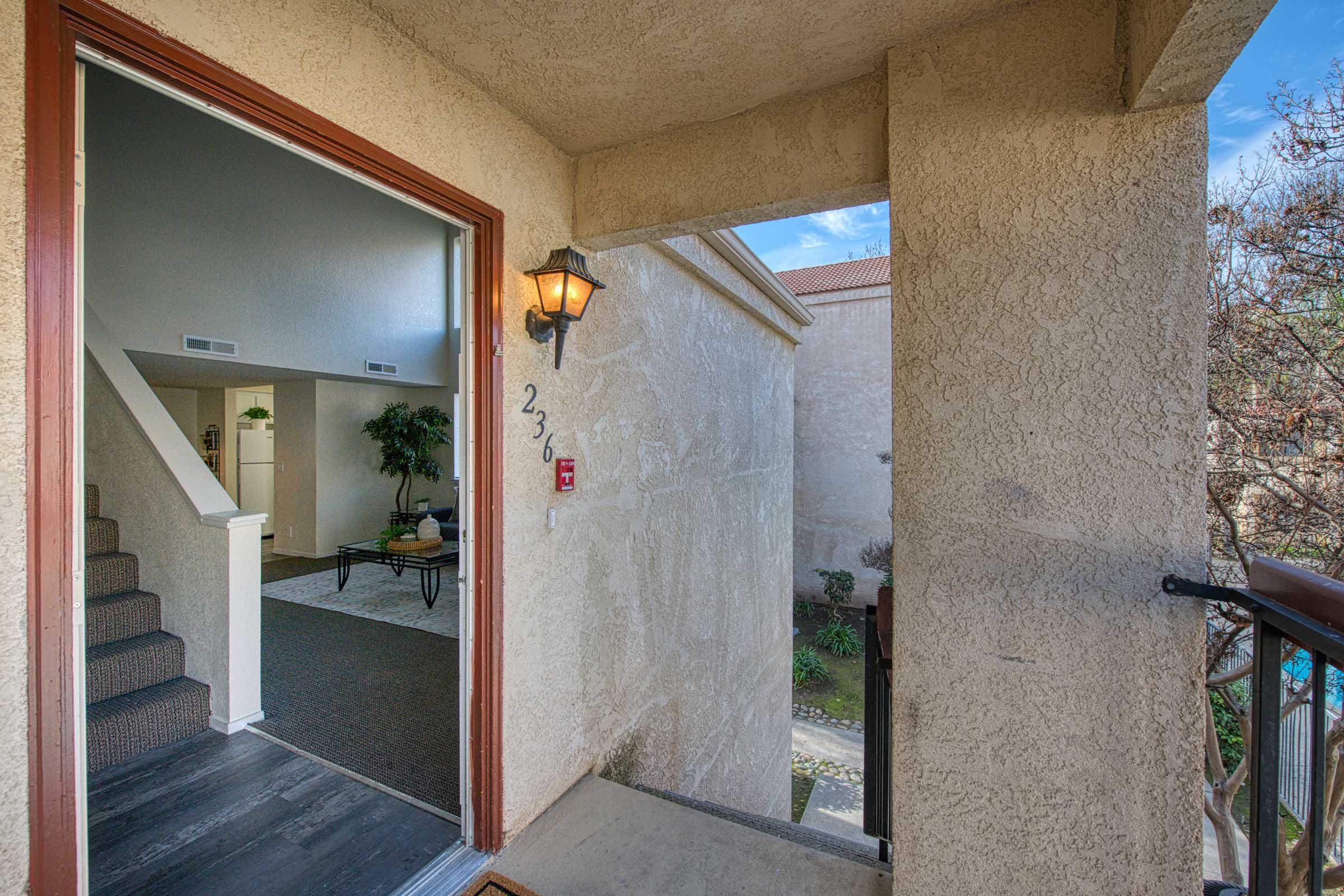 View from a balcony looking towards an open doorway. A staircase is visible leading to the upper level inside. The exterior wall is textured, with a light fixture beside the door. The entrance is numbered 236, and there are plants visible in the background.