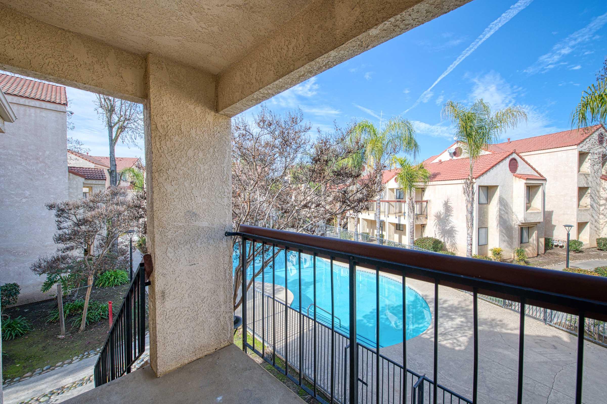 View from a balcony overlooking a swimming pool surrounded by palm trees and landscaped gardens. The scene features a clear blue sky with a few clouds and buildings in the background, creating a serene and inviting atmosphere.