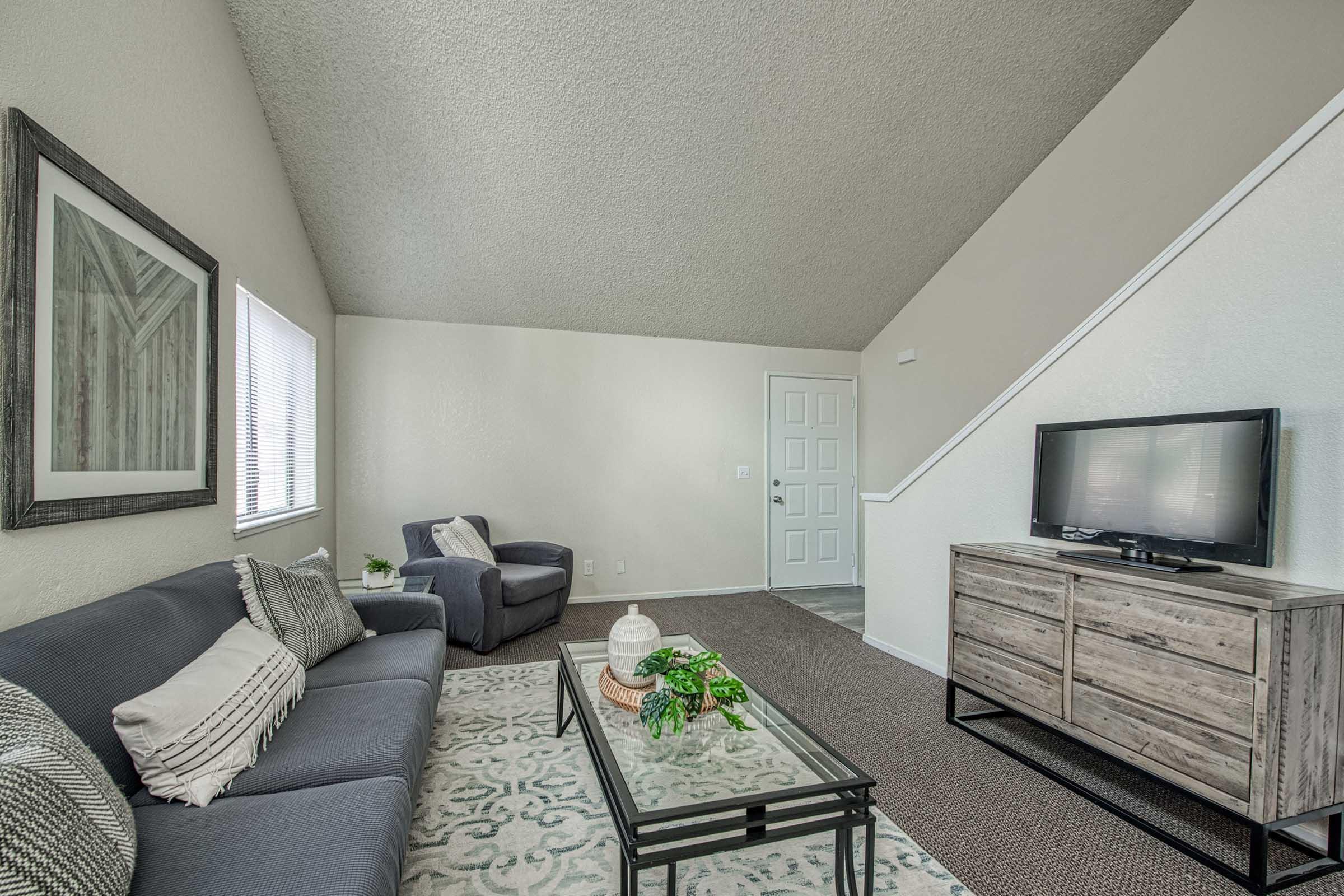 A cozy living room featuring a gray sofa with decorative pillows, a dark accent chair, a glass coffee table with green decor, a wooden media console with a TV, and a staircase in the background. Natural light pours in through windows, and the walls are painted in soft neutral tones.