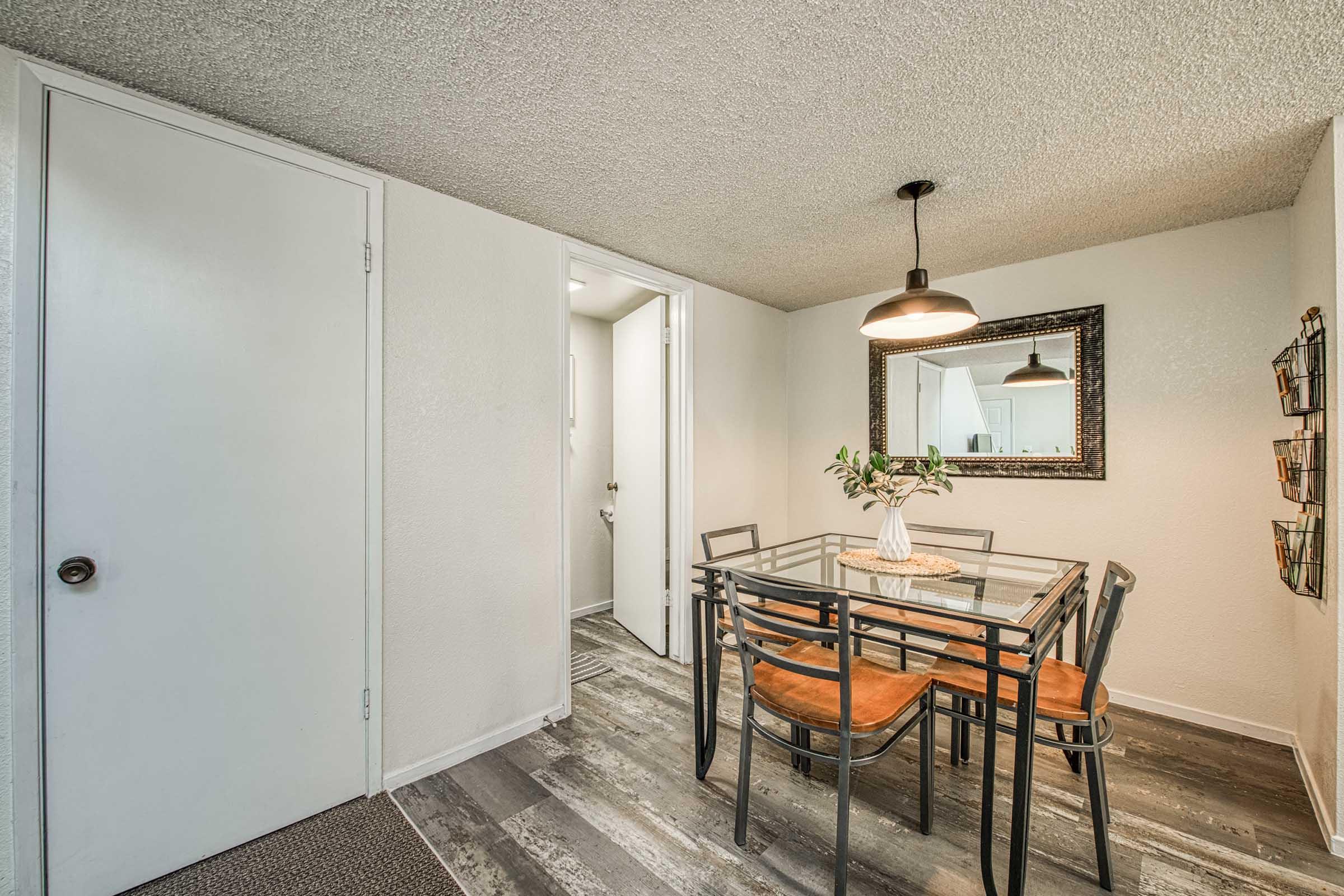 A well-lit dining area featuring a glass-top table with four chairs, a vase of flowers at the center, a large wall mirror, and decorative wall shelves. The space has neutral-colored walls and modern flooring, with a door leading to another room visible in the background.