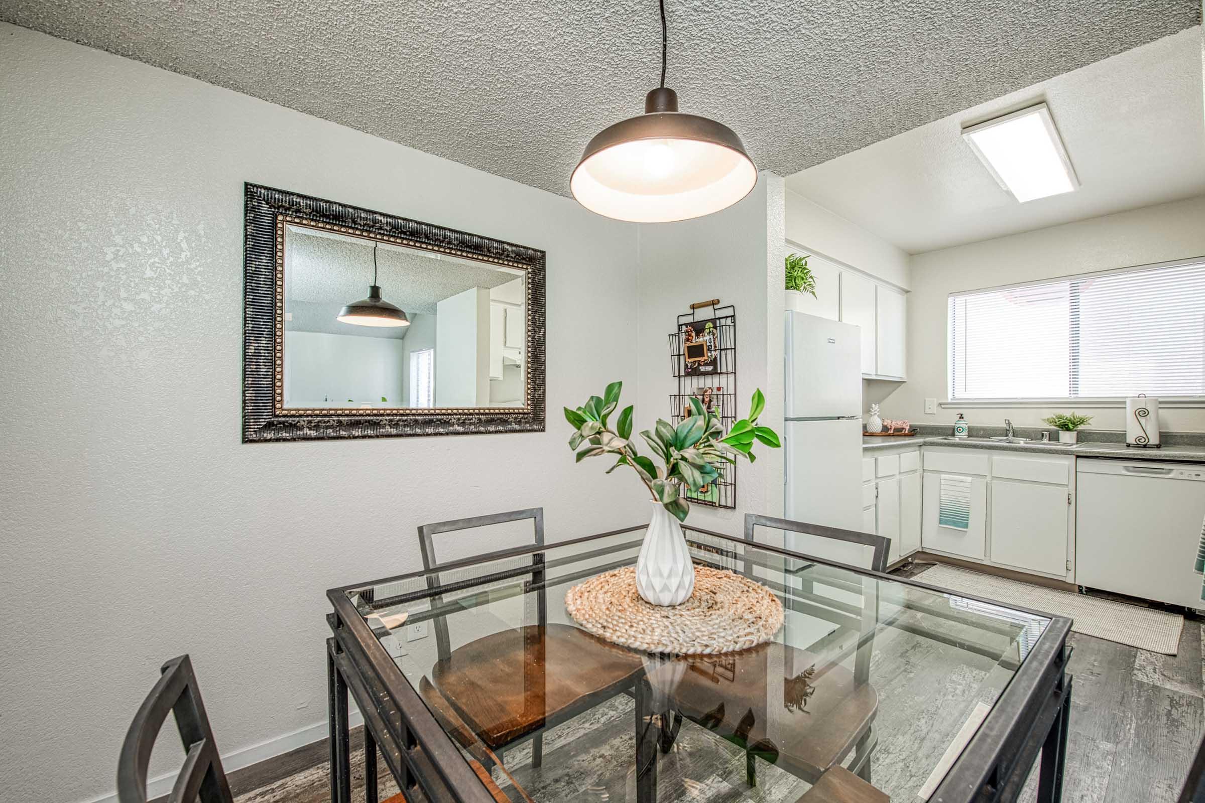 A modern kitchen and dining area featuring a glass-top dining table with black metal chairs, a decorative vase with greenery in the center, a large mirror on the wall, and a well-lit kitchen in the background with white cabinets and appliances. Natural light streams in through a window.