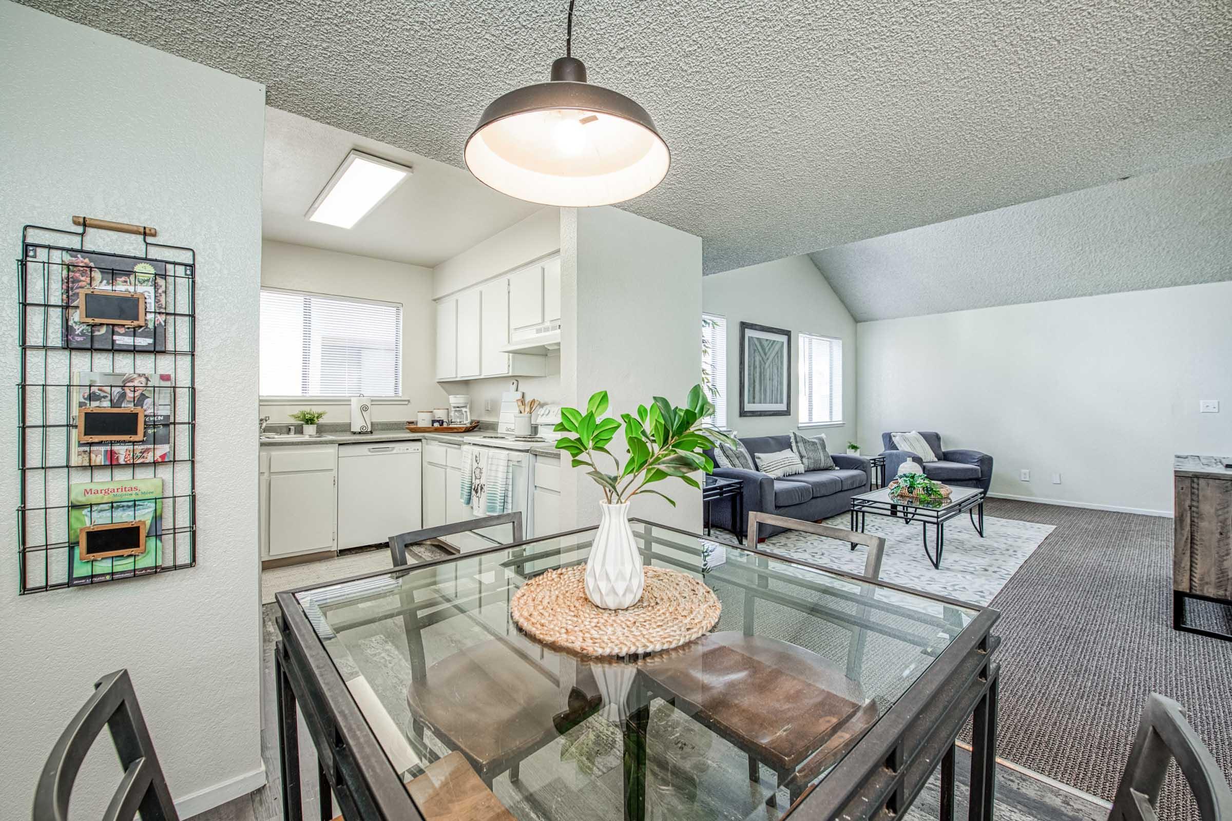 A modern living space featuring a glass dining table with a centerpiece of green leaves, surrounded by black chairs. In the background, a kitchen area with white cabinets and appliances is visible. A cozy living room with a gray sofa and decorative rug can be seen to the right, illuminated by natural light.