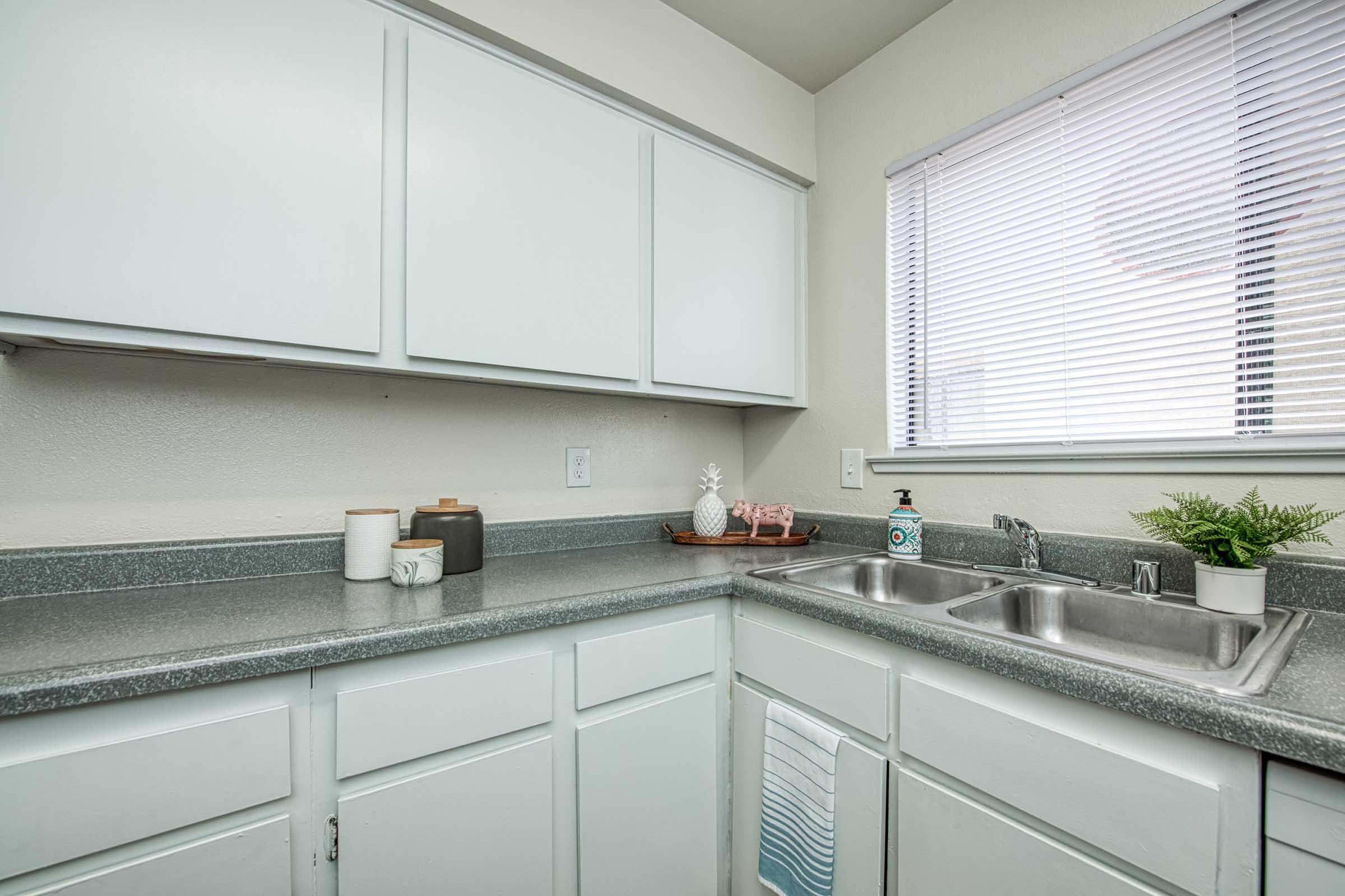 A modern kitchen corner featuring a gray countertop, white cabinetry, and a window with blinds. There are two stainless steel sinks, a small potted plant, and decorative items including a pineapple figurine and bottles on a tray. The overall color scheme is light and neutral.
