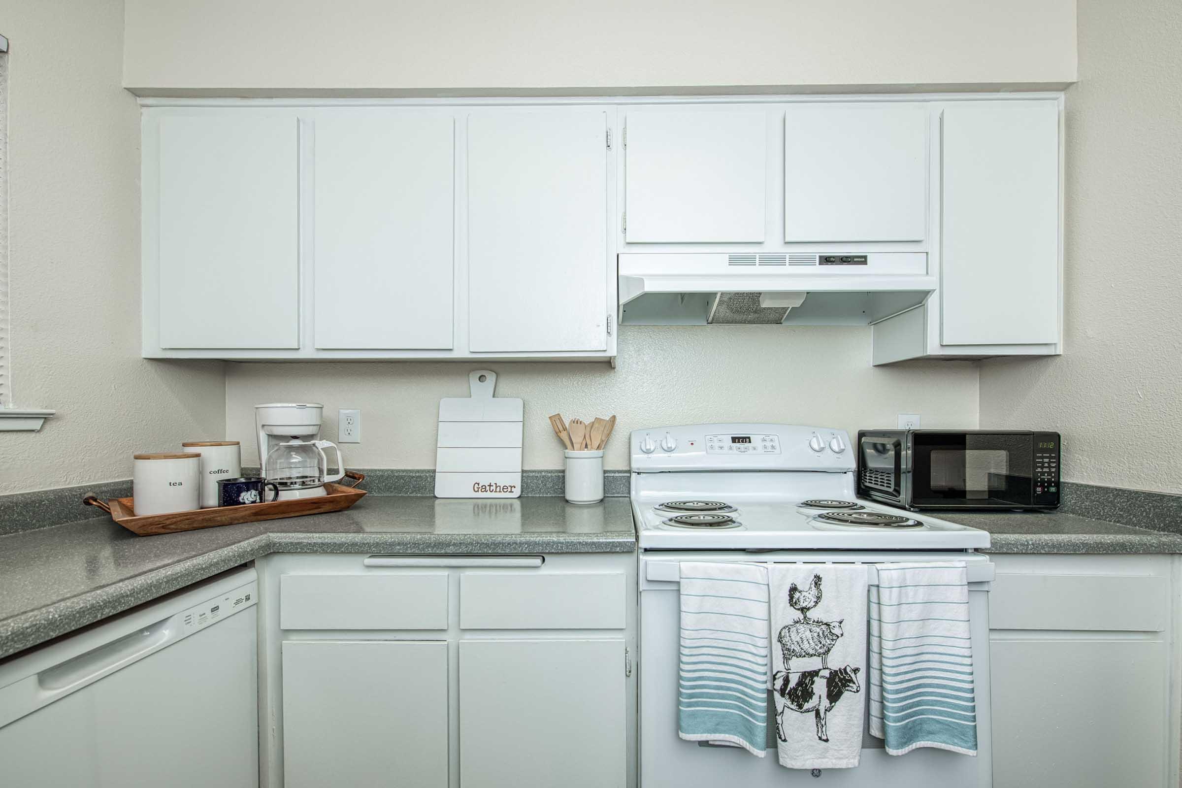 A bright, modern kitchen featuring white cabinets and a grey countertop. The stove with two burners is flanked by a microwave and a coffee maker. A dish towel with a decorative design hangs on the stove. A wooden tray holds coffee supplies, while utensil holders and baking tools are visible on the counter.