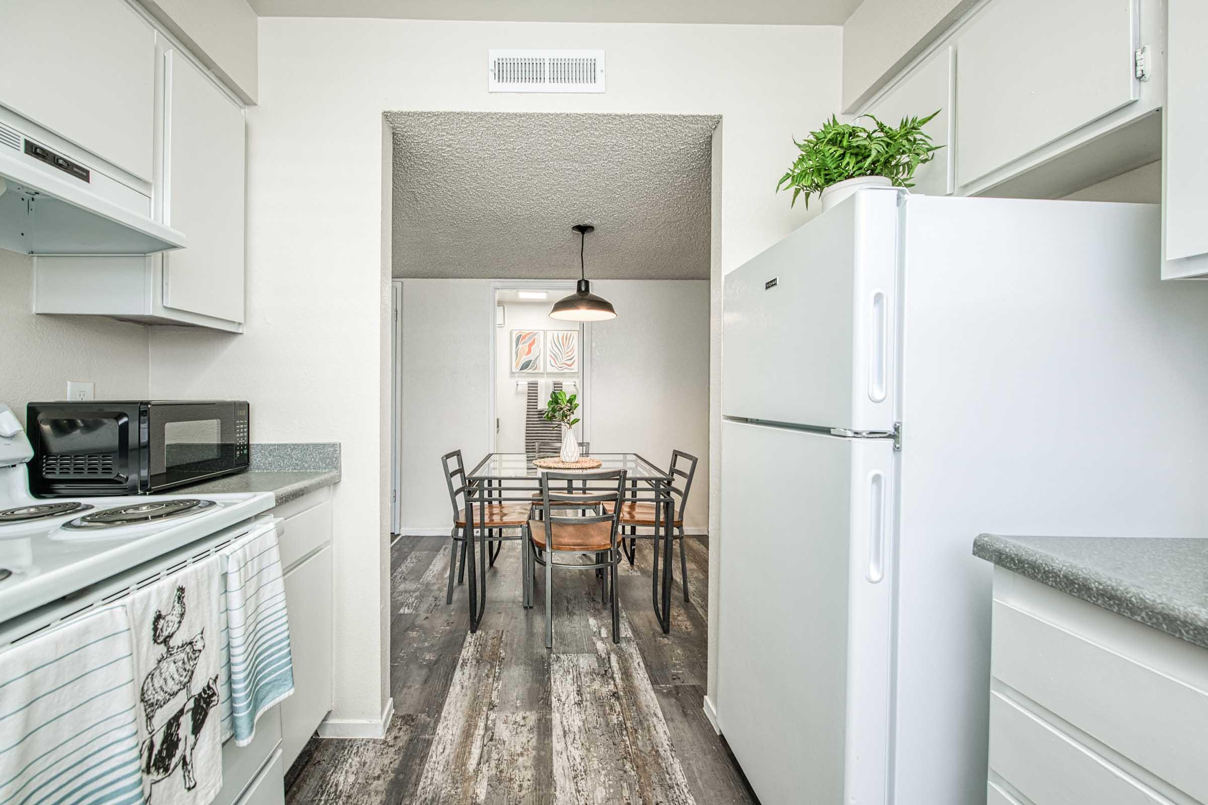 Bright and modern kitchen with a white refrigerator, microwave, and stove. Features a dining area with a round table and black chairs, illuminated by a pendant light. Light-colored cabinetry and grey wood-style flooring create an inviting atmosphere. A potted plant adds a touch of greenery.
