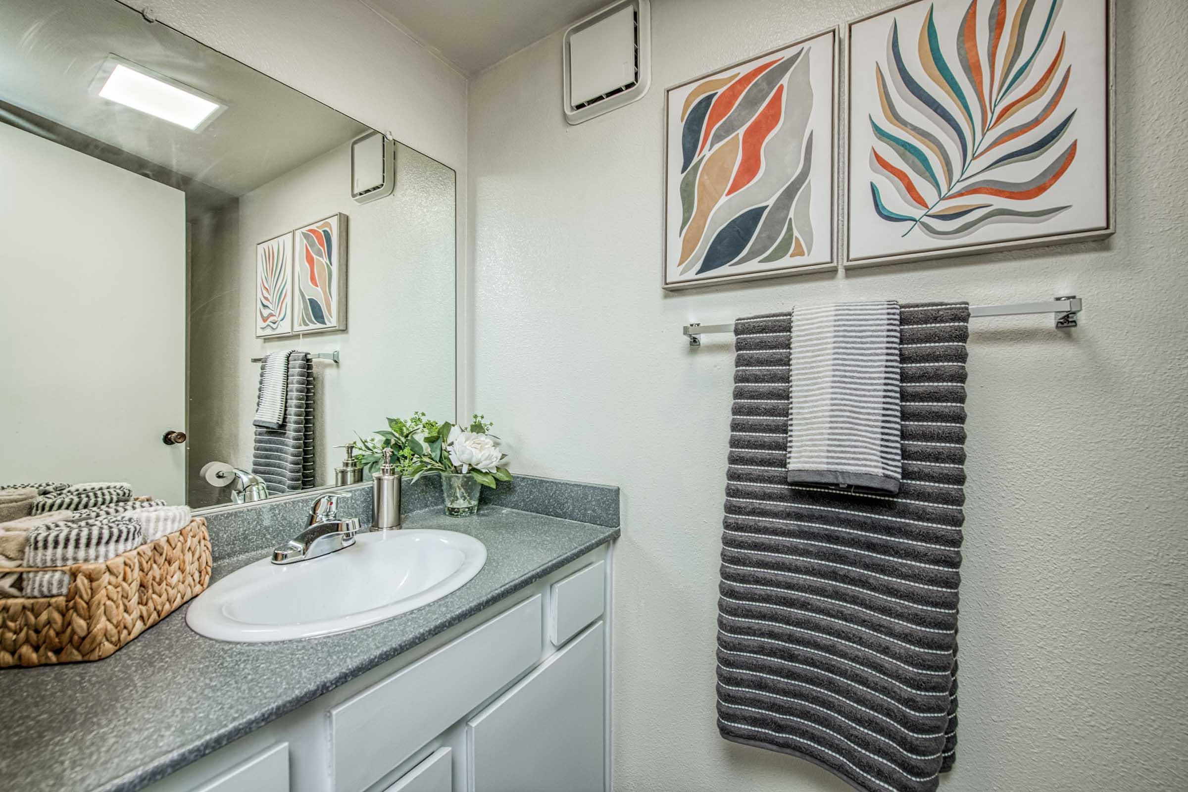 A clean and modern bathroom featuring a sink with a mirror above, decorative wall art, gray striped towels hanging on a rack, and a wicker basket containing toiletries. The countertop is sleek with a small vase of flowers, creating a fresh and inviting atmosphere.