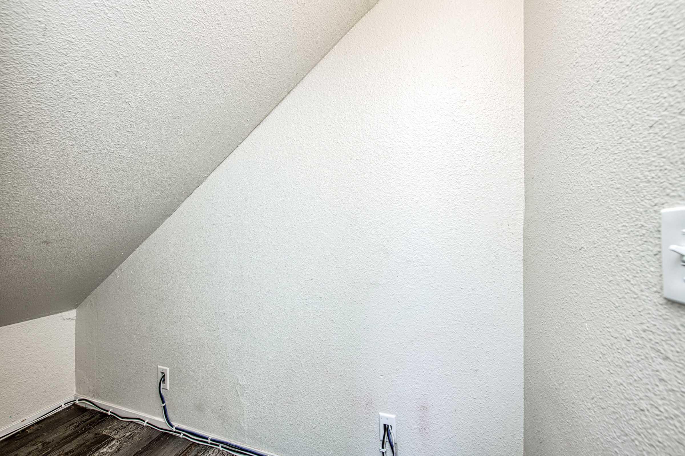 A small, empty corner of a room with a sloped ceiling. The walls are painted white, and the floor has dark wood-like flooring. There are electrical outlets on the wall, but no furniture or decorations are present, giving the space a minimalistic appearance.