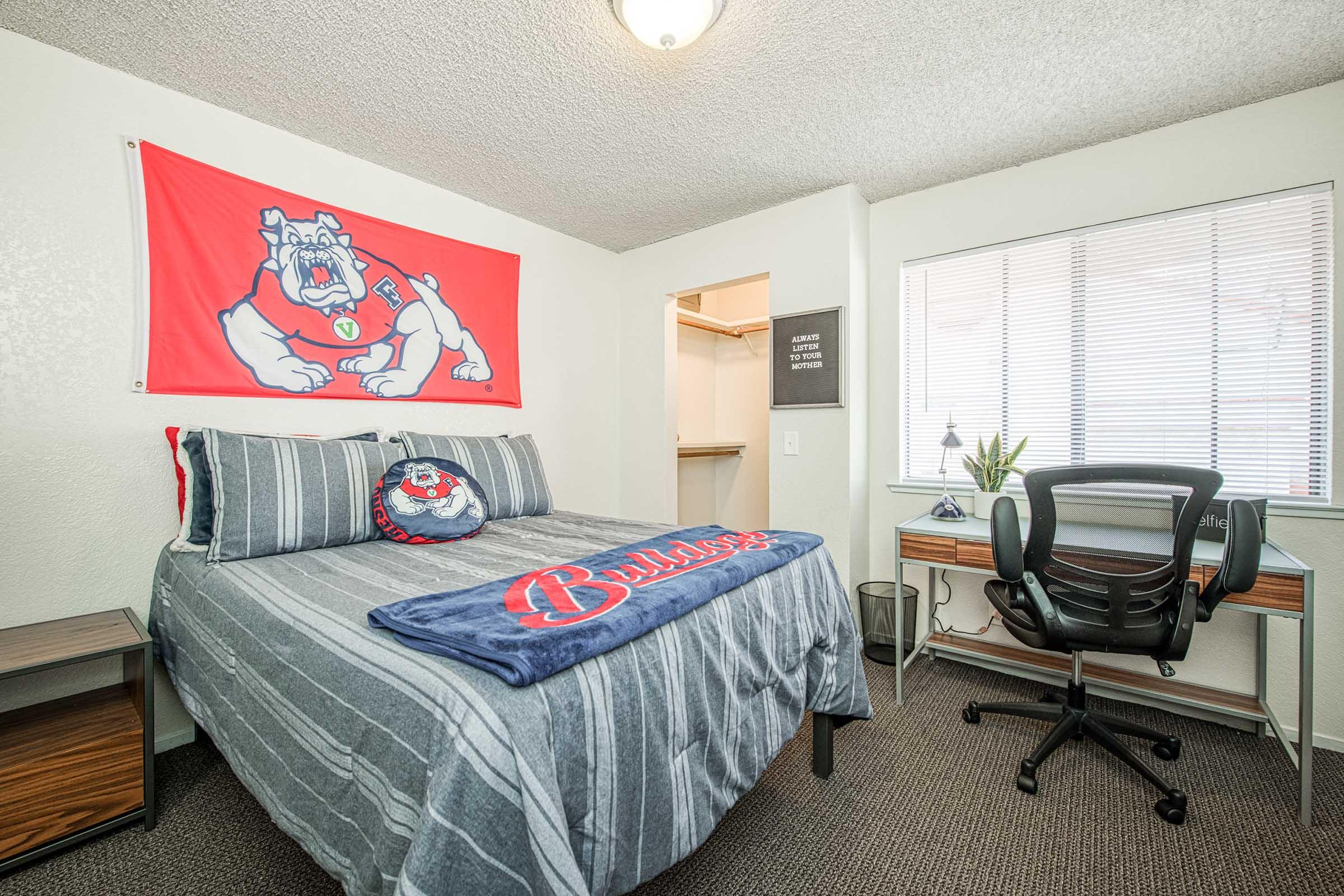 A cozy bedroom featuring a large bed with gray striped bedding and a Bulldogs-themed blanket. There's a flag of the Bulldogs hanging on the wall, a desk with a chair, and a small plant near the window. The room has neutral walls and a carpeted floor, creating a welcoming and functional study space.