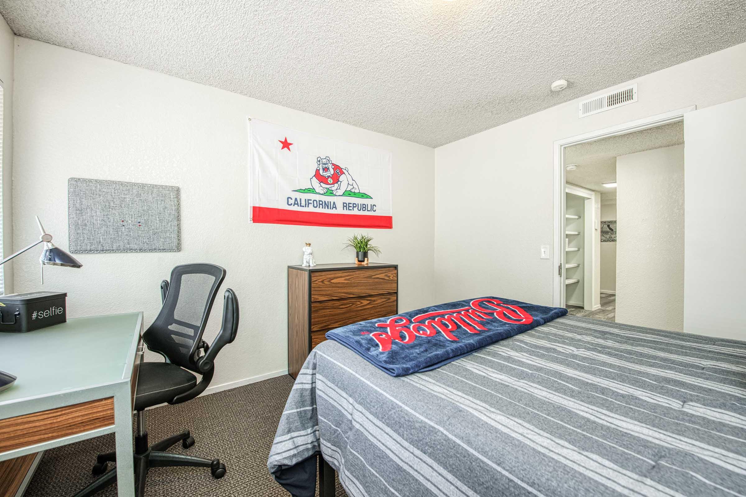 A bedroom featuring a California Republic flag hanging on the wall. There's a bed with a blue and gray striped blanket, a wooden dresser, a desk with a chair, and a wall-mounted bulletin board. The room has a neutral color scheme and minimal decor, giving it a cozy and organized feel.