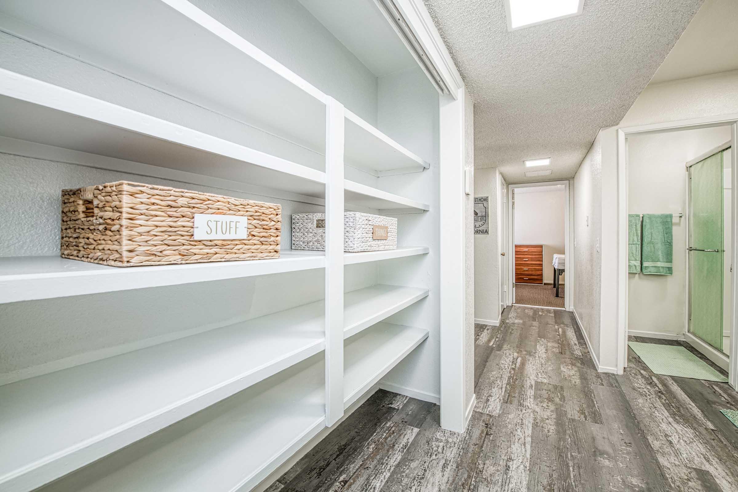A spacious, well-organized bathroom with white shelves displaying decorative storage baskets labeled "STUFF." The floor features a modern wood-like design, and a glass-enclosed shower is visible in the background, along with a hallway leading to other rooms. Soft, neutral colors enhance the inviting atmosphere.