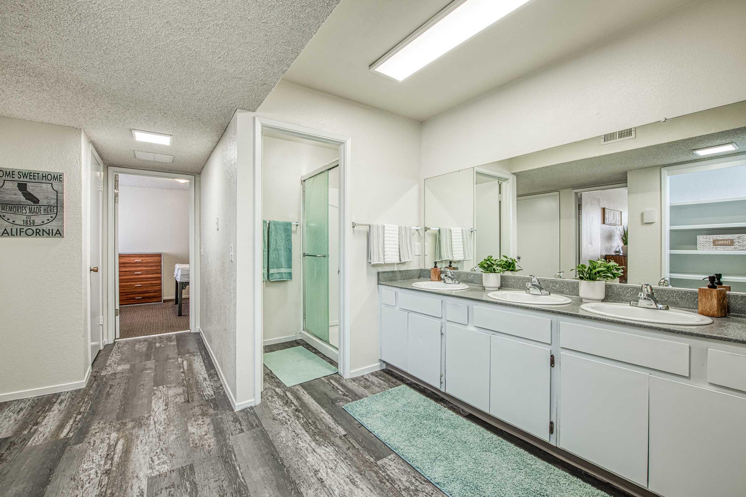 Modern bathroom featuring dual sinks with a long mirror, light wood cabinetry, and a glass shower. The decor includes potted plants and towels. A light-colored floor complements the bright, open space, with an adjacent room visible in the background.