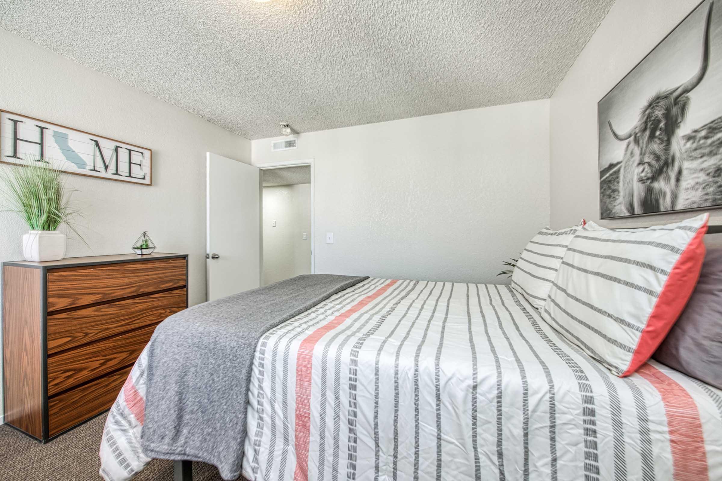 A cozy bedroom featuring a neatly made bed with striped bedding, a wooden dresser, and a plant. A large wall art piece of a bull hangs above the bed. The room has light-colored walls and a door leading to another area, creating a warm and inviting atmosphere.