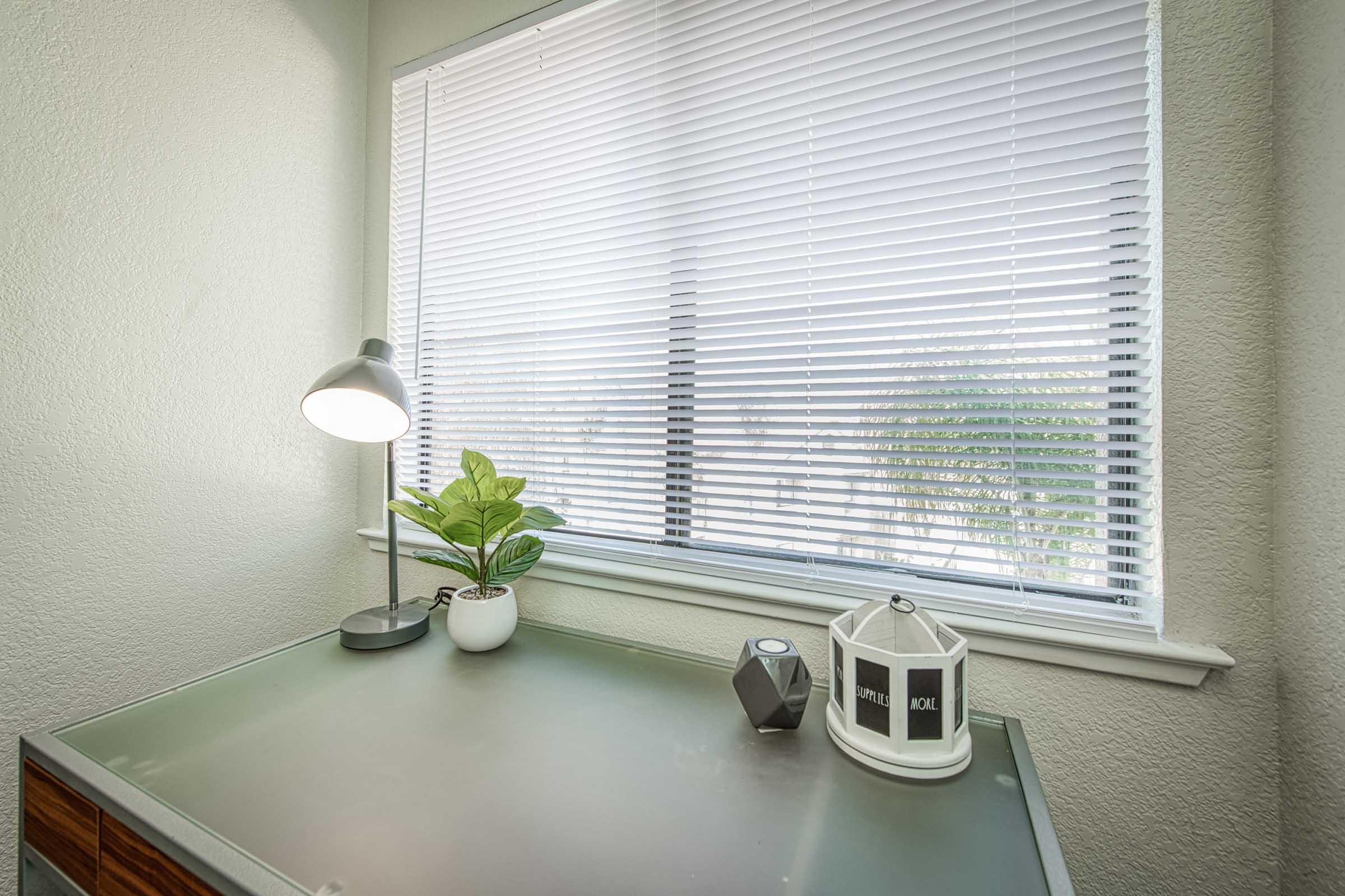 A modern workspace featuring a sleek desk with a glass surface, a small white potted plant, a minimalist lamp, and decorative items including a geometric object and a lantern. Natural light filters through white blinds, illuminating the area and providing a serene atmosphere.