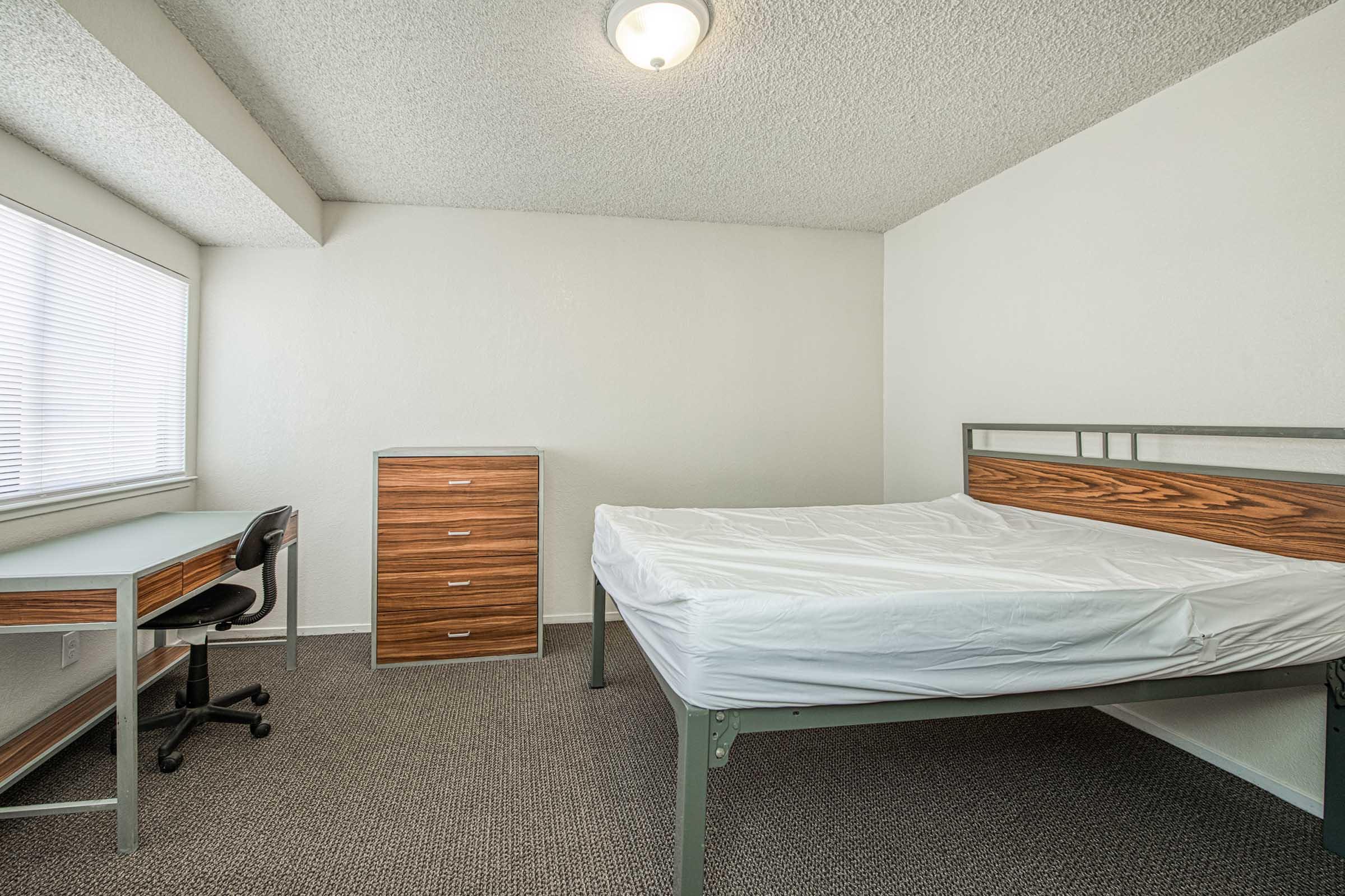 A simple bedroom featuring a metal bed frame with a mattress covered in a white fitted sheet, a wooden dresser with five drawers, and a desk with an office chair. The room has a large window allowing natural light and is decorated in neutral tones with carpeted flooring.