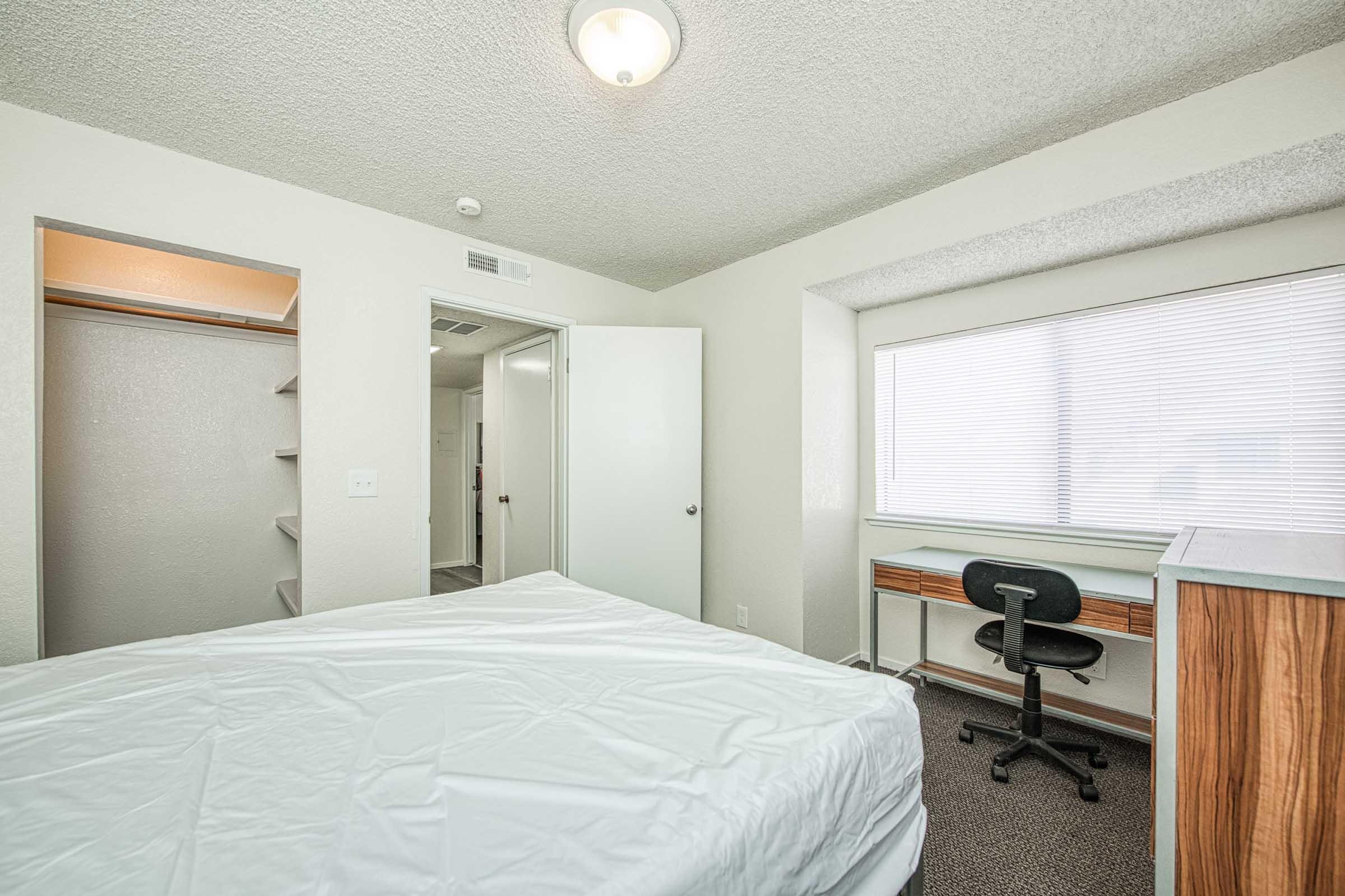 A cozy bedroom featuring a bed with a white mattress, a desk with a chair by a window, and a closet. The room has textured walls, a ceiling light, and light-colored furnishings. Natural light filters through the window, creating a welcoming atmosphere.