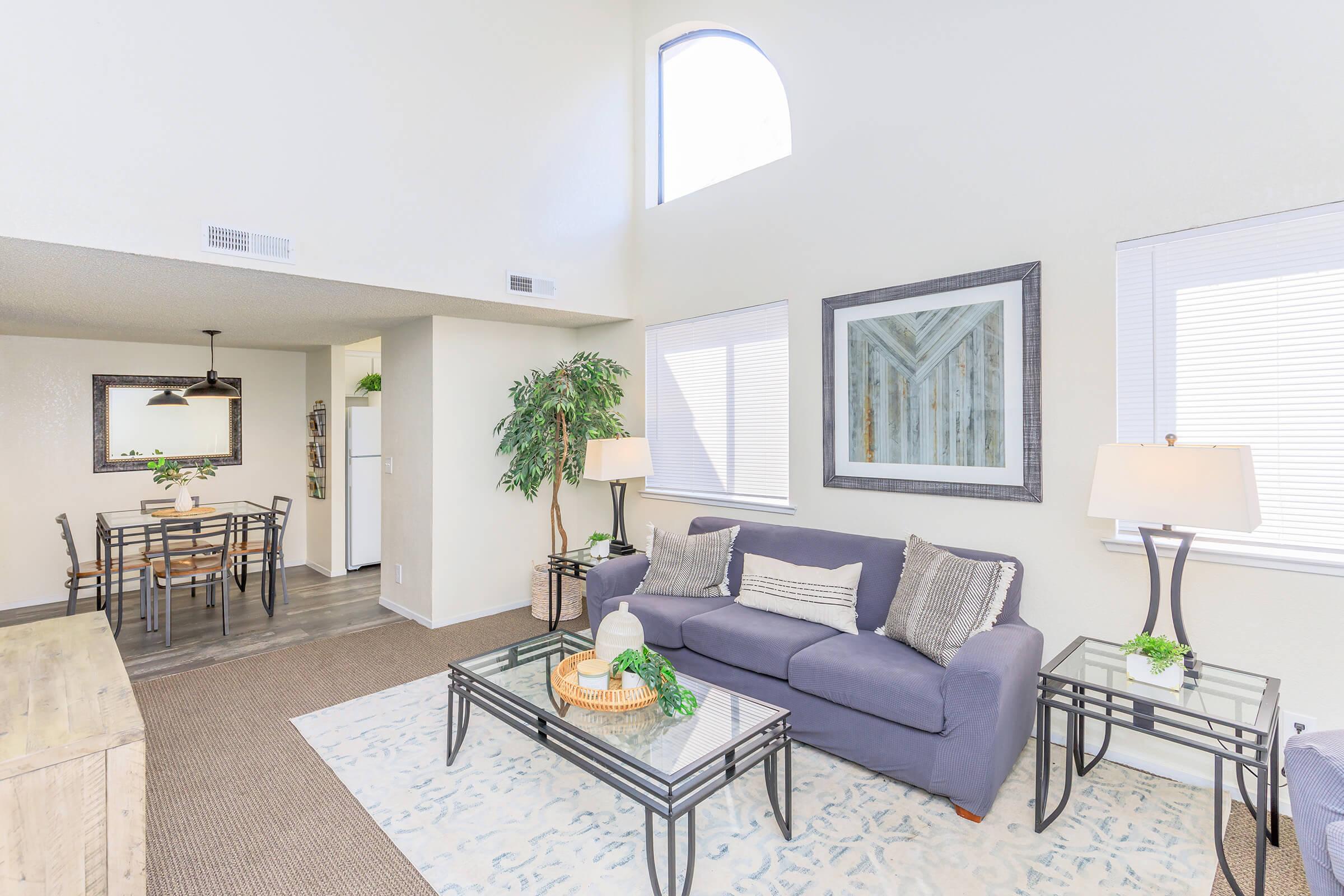 A cozy living room featuring a blue sofa with decorative pillows, a coffee table with a plant, and two lamps. A dining area with a wooden table and chairs is visible in the background, along with a kitchen area. Large windows allow natural light to fill the space, creating a bright and inviting atmosphere.