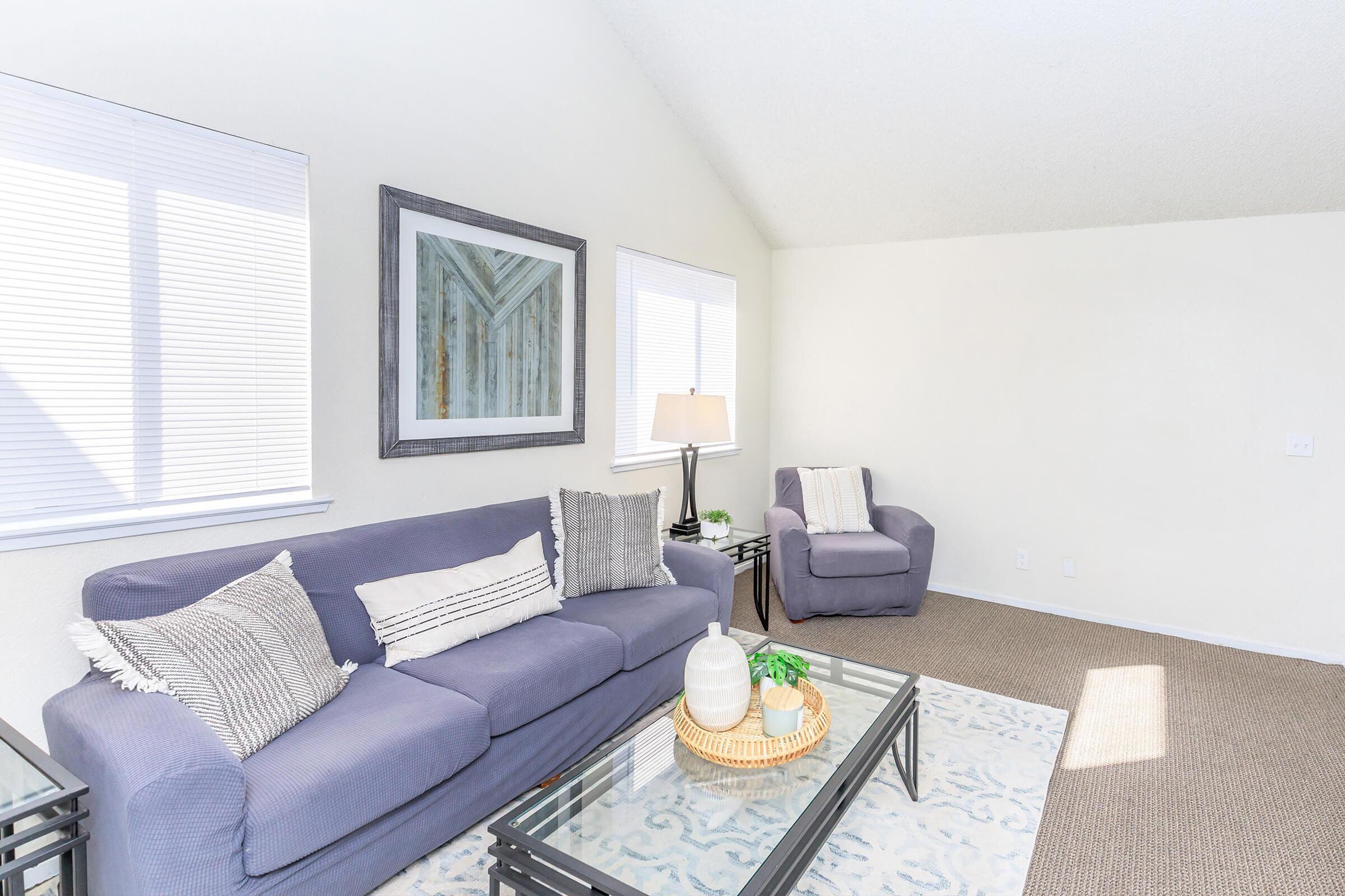 A bright and airy living room featuring a light gray sofa with decorative pillows, a matching armchair, a glass coffee table with a plant, and artwork on the wall. Natural light floods in through two windows with blinds, creating a welcoming atmosphere.