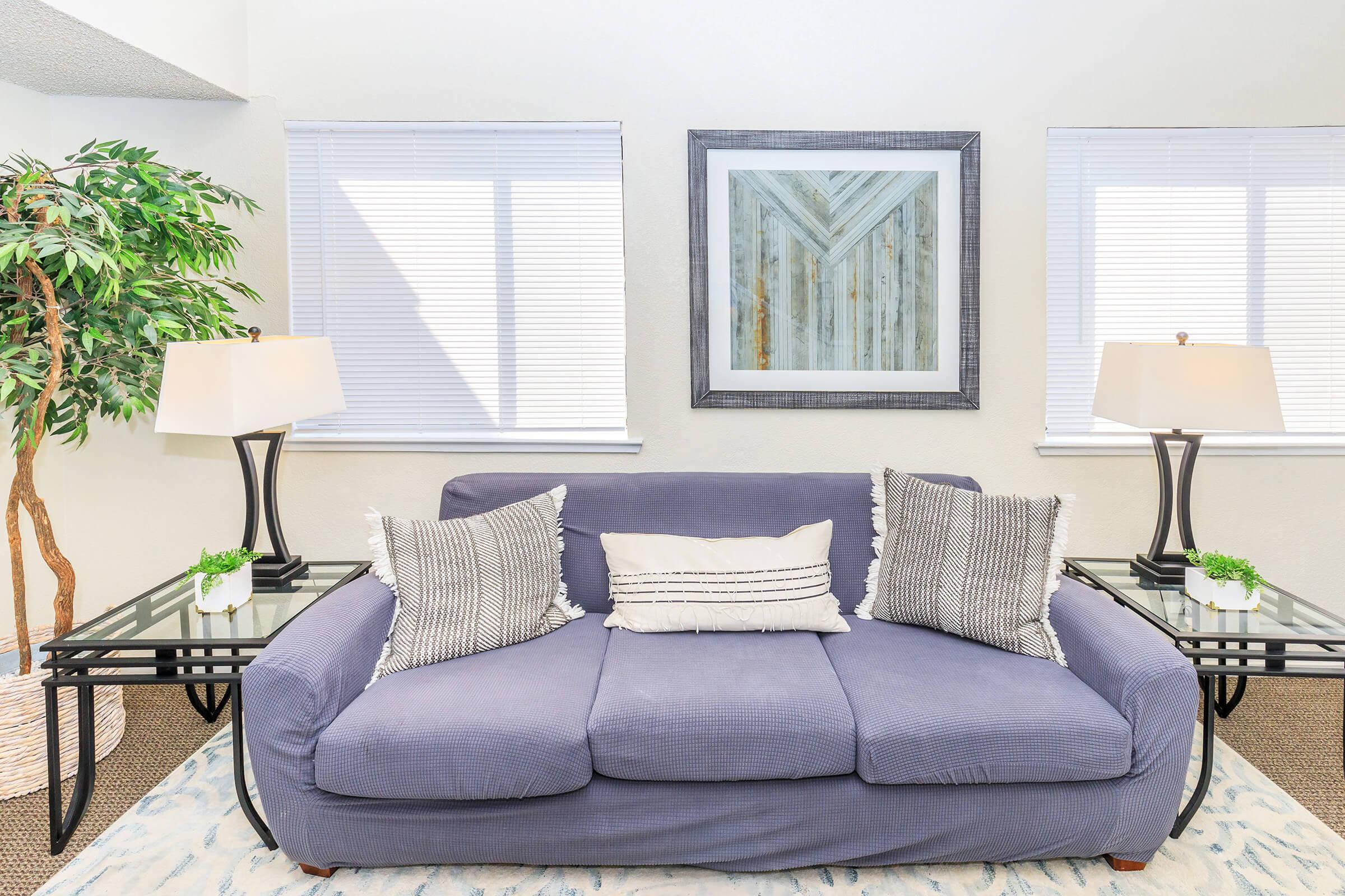 A cozy living room featuring a purple sofa with two textured pillows, flanked by black glass side tables with small potted plants. Two elegant table lamps provide soft lighting, while light curtains cover the windows. A framed abstract artwork hangs on the wall, enhancing the inviting atmosphere.