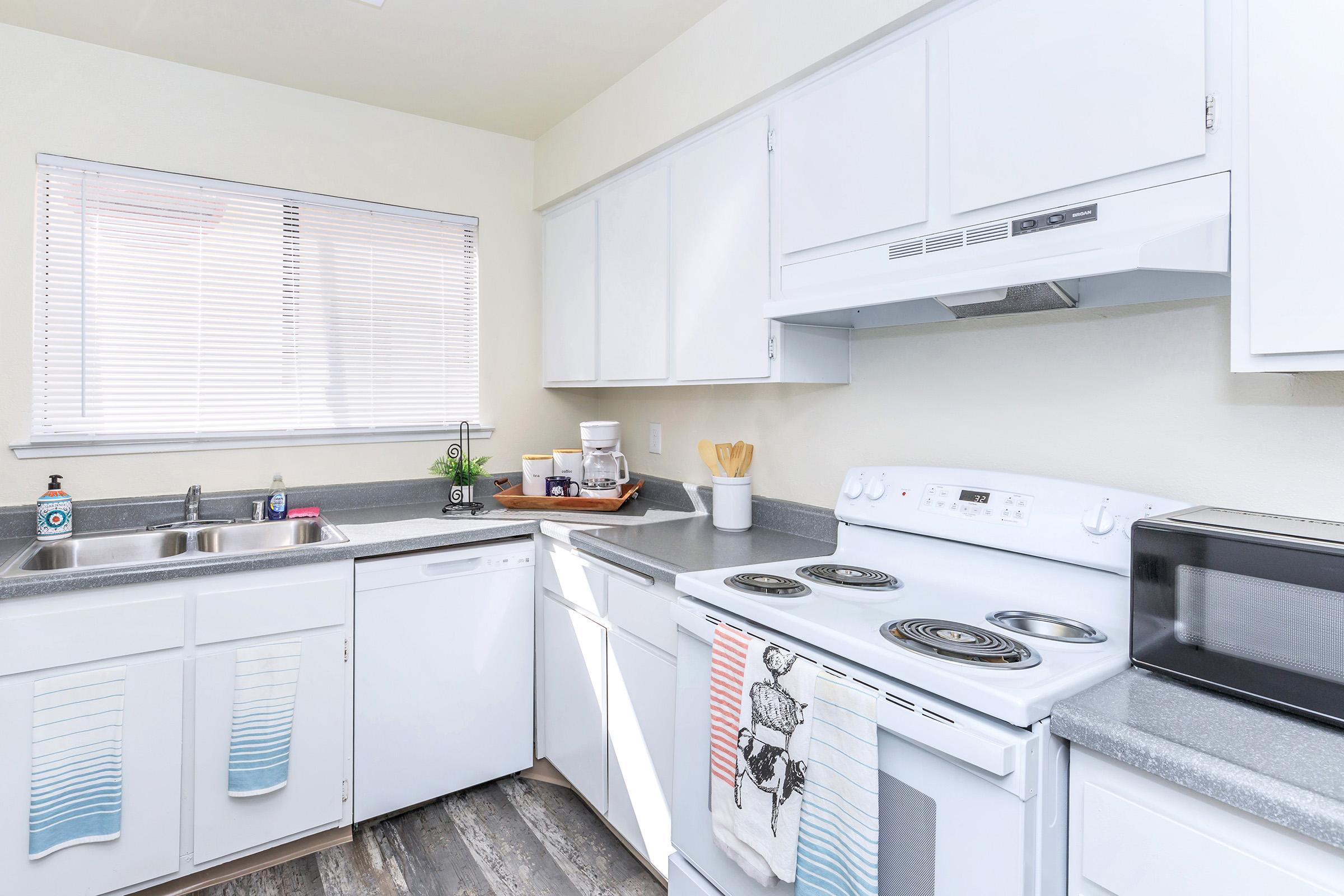 Bright and modern kitchen featuring white cabinets, a gray countertop, and stainless steel appliances. The kitchen includes a sink, dishwasher, stove, and microwave, with a window providing natural light. A small plant and kitchen utensils are placed on the counter, creating an inviting atmosphere.