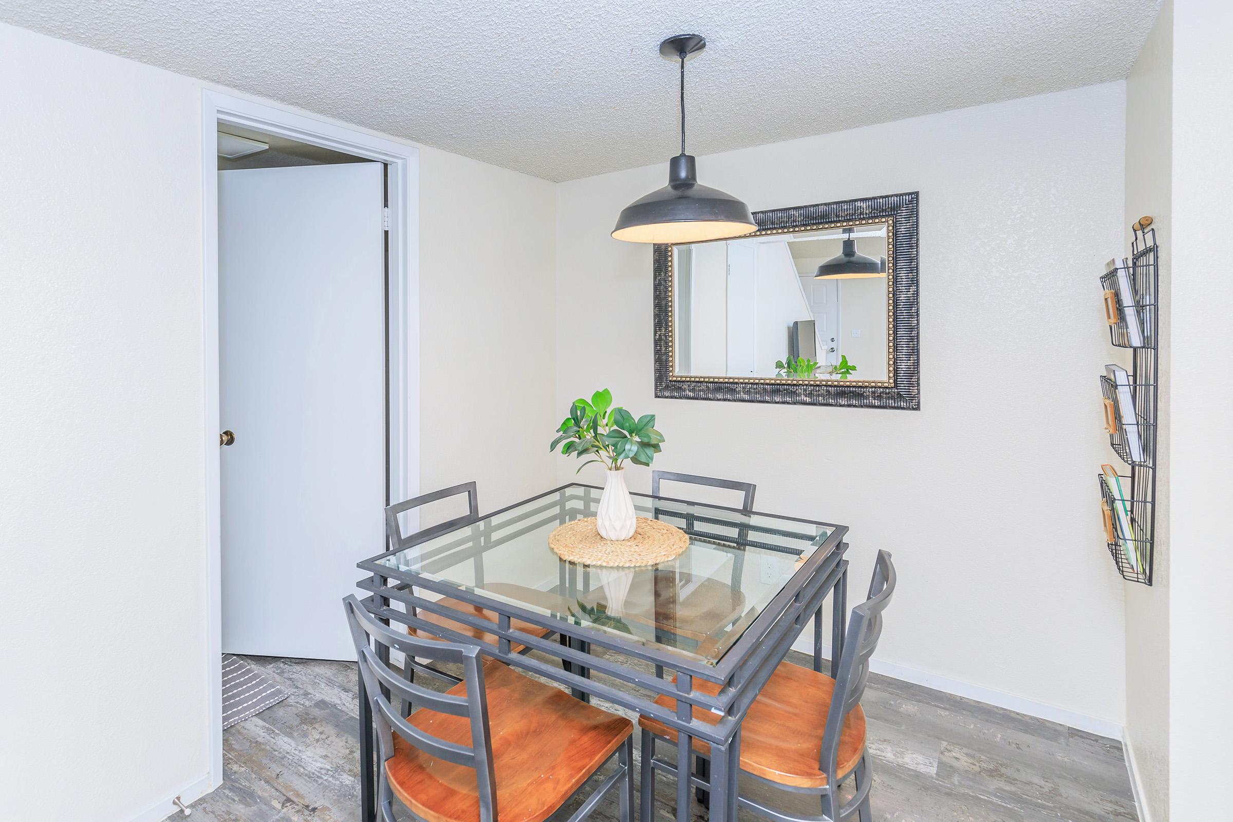 A small dining area featuring a glass-top table with four black metal chairs and wooden seats. A simple vase with a plant is at the center of the table. A large mirror is hanging on the wall, enhancing the space. The walls are light-colored, and the flooring is wood-like.