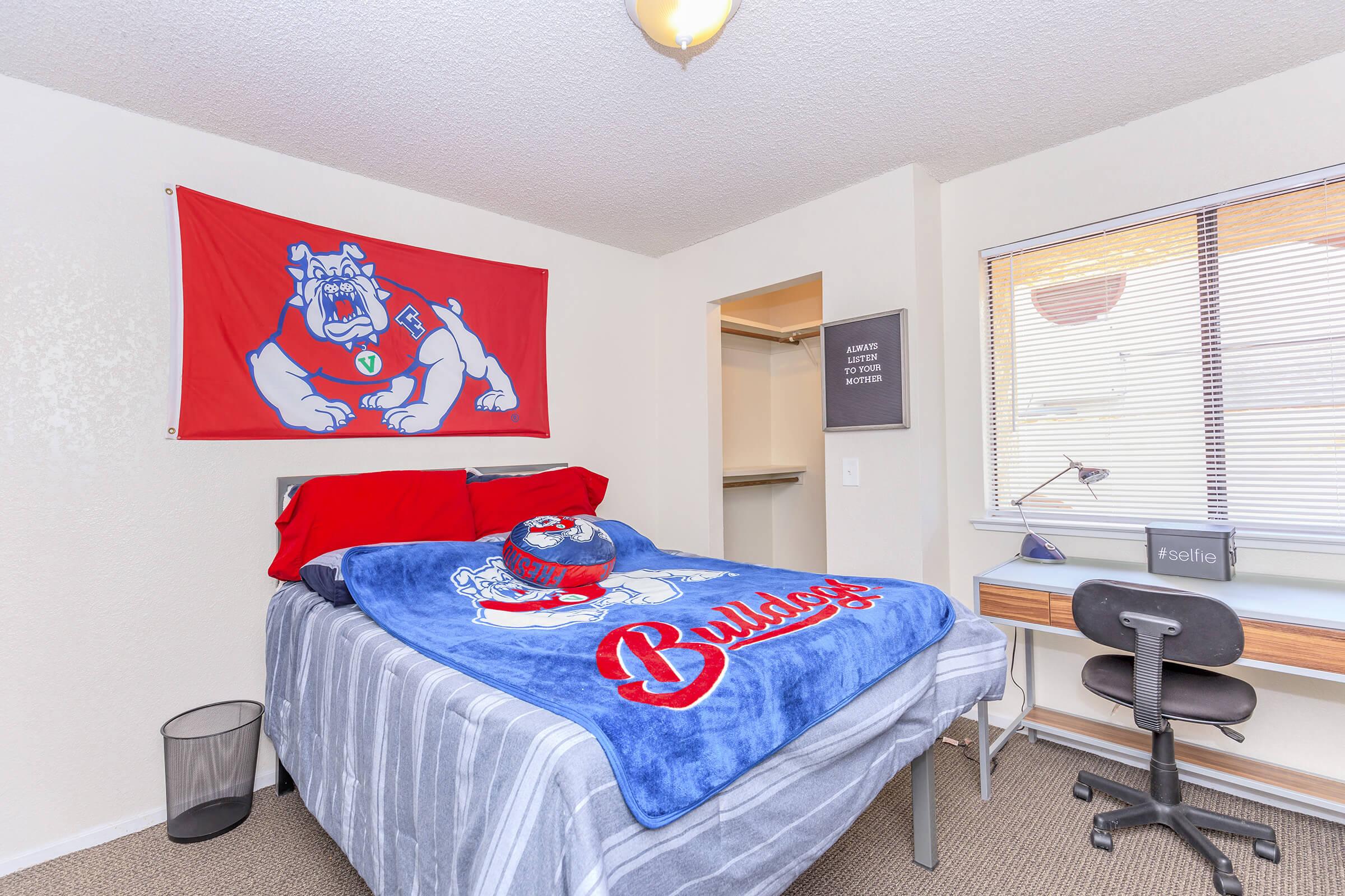 A cozy bedroom featuring a bed with a Bulldog-themed comforter and decorative pillows. A red Bulldog flag hangs on the wall. There's a small desk with a chair by a window, a wastebasket in the corner, and a closet space visible. Natural light brightens the room, creating a welcoming atmosphere.