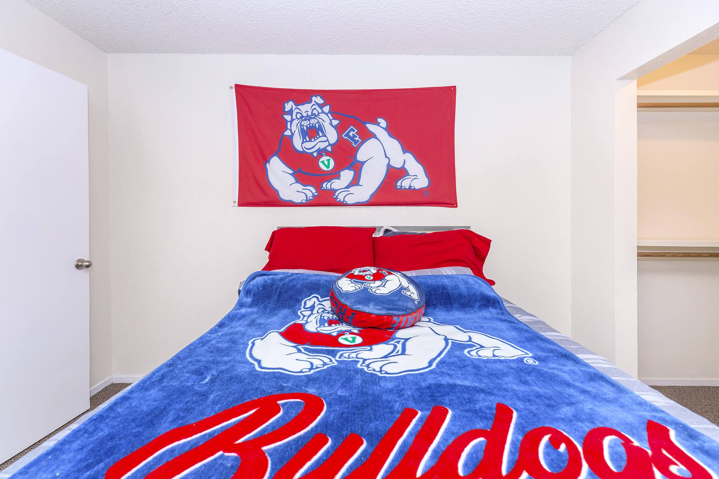 A cozy bedroom featuring a red and blue color scheme. The bed is dressed with a Bulldog-themed blanket and pillow, and there's a large flag of a Bulldog mascot hanging on the wall. The room has a simple design with white walls and a closet visible in the background.