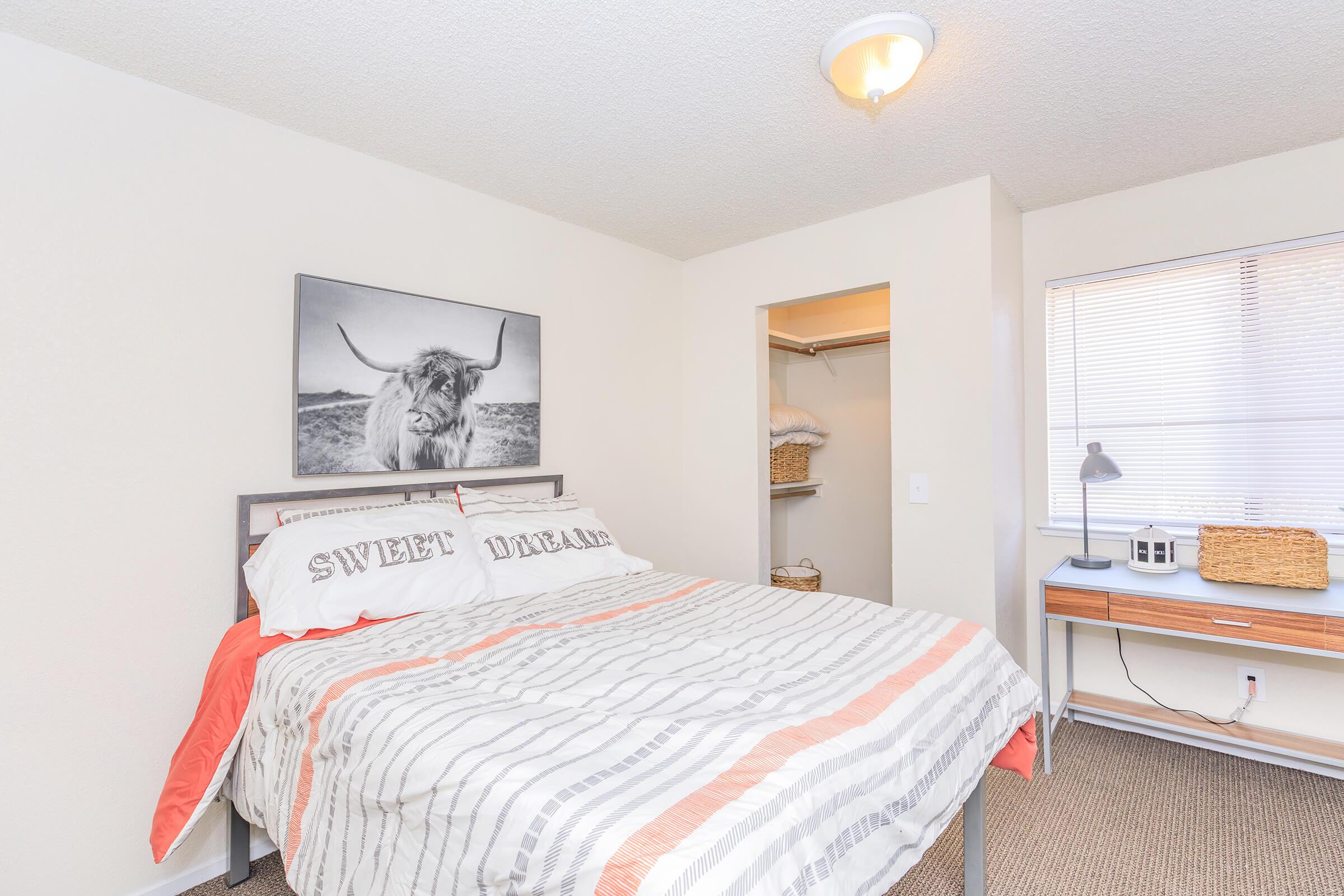 A cozy bedroom featuring a bed with striped bedding and "SWEET DREAMS" pillows. The room has a large window with light curtains, a small desk with a lamp, and an open closet displaying clean towels. A black-and-white photo of a Highland cow hangs above the bed.