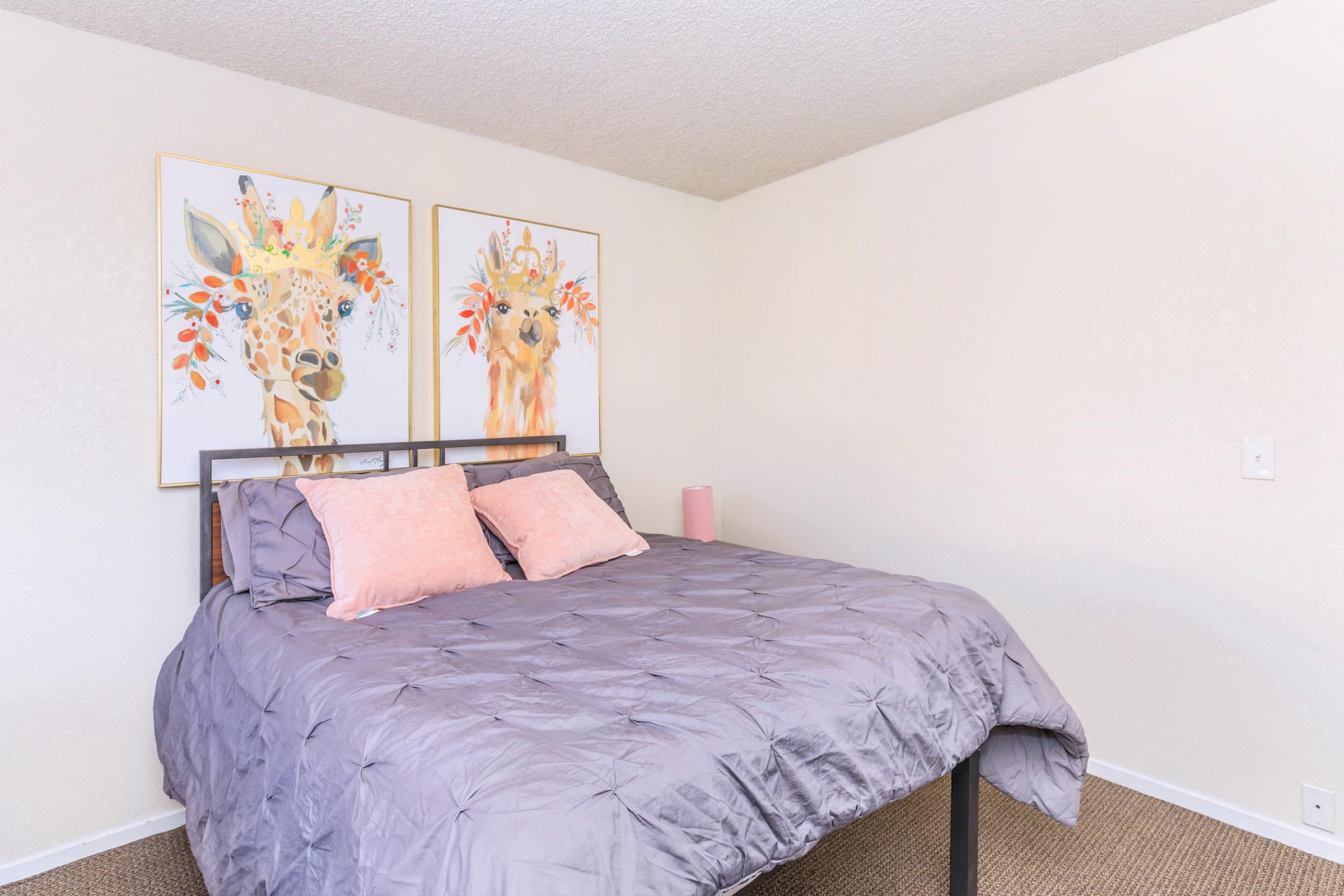 A cozy bedroom featuring a queen-sized bed with a gray comforter and soft pink pillows. Two vibrant animal-themed wall art pieces adorn the walls, depicting giraffes decorated with floral accents. The room has light-colored walls and a minimalistic design with a lamp beside the bed.