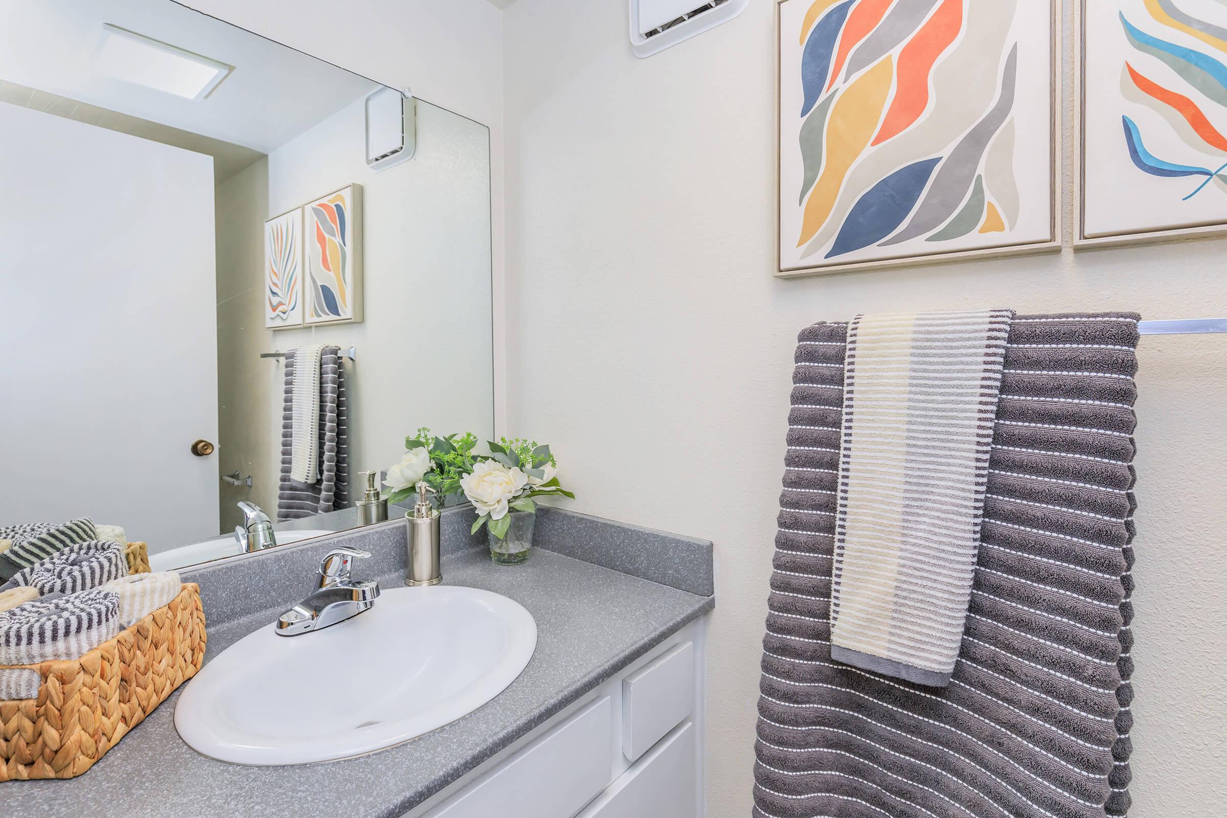 A modern bathroom with a gray countertop, a silver faucet, and a mirror reflecting colorful artwork on the wall. A neatly arranged towel hangs on a rack, and there's a woven basket containing rolled towels and a small vase with white flowers.