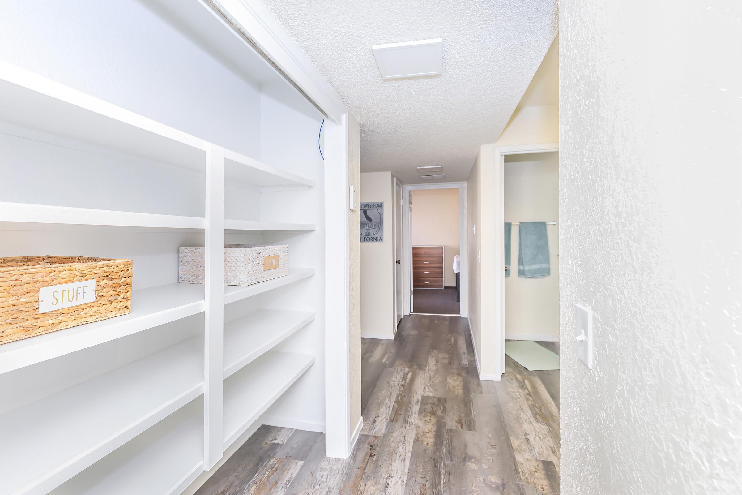 A well-lit hallway featuring a built-in shelving unit on the left with wicker storage baskets labeled "STUFF." The floor has a modern wood-like finish, and there are doors leading to other rooms, including a bathroom visible at the end of the hallway. The walls are painted in neutral colors.