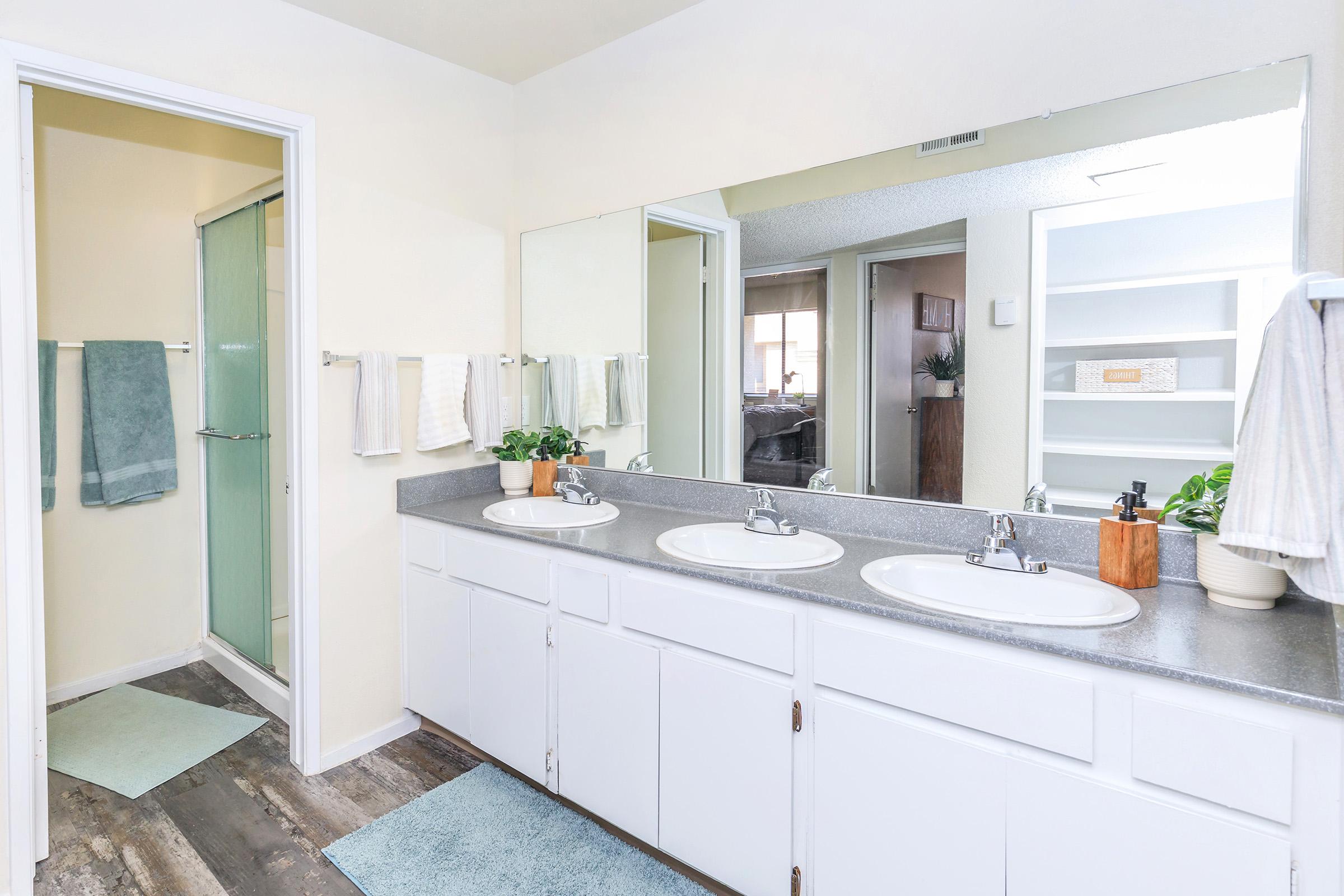 A modern bathroom featuring a double sink vanity with two sinks, a large mirror above, and a shower area to the left. The decor includes soft blue towels, clean white cabinetry, and light-colored walls, creating a fresh and inviting atmosphere.