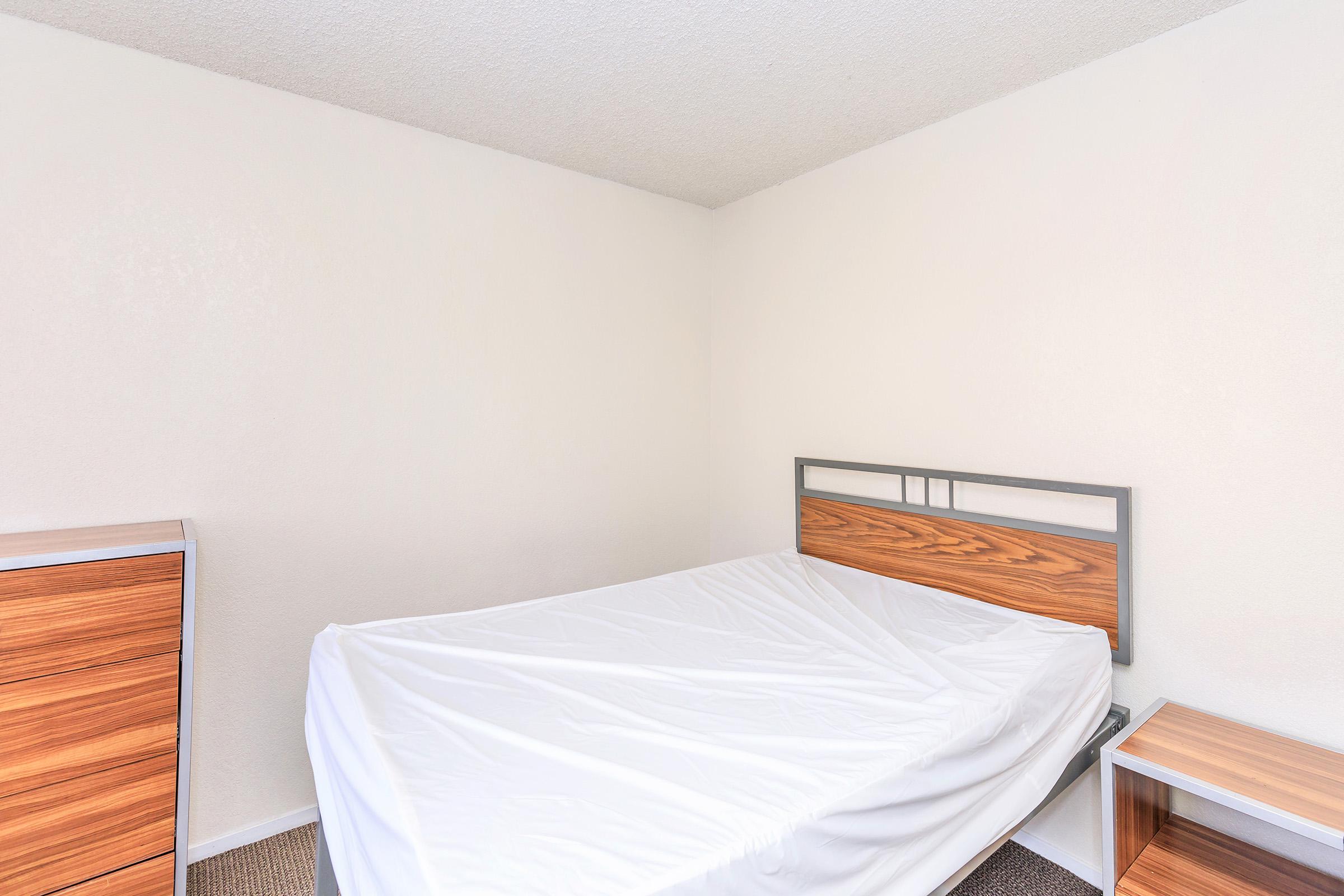 A simple, unadorned bedroom featuring a bed with a white mattress cover, a wooden headboard, and two wooden nightstands. The walls are plain and white, and the floor has a light carpet. The room appears minimalistic and uncluttered.