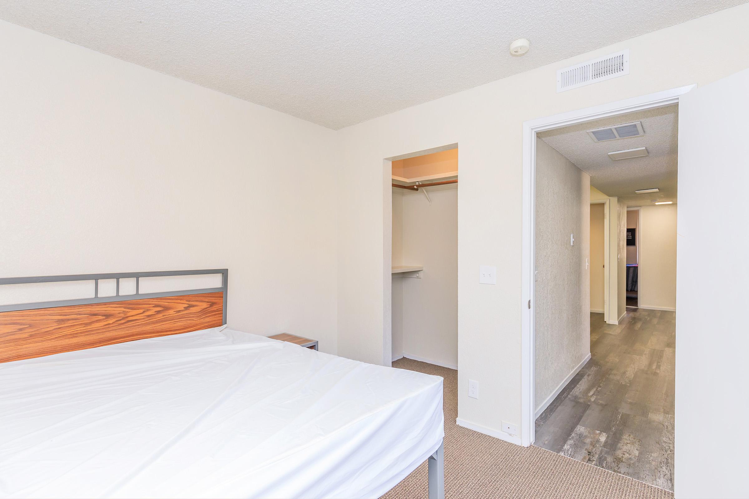 A lightly furnished bedroom featuring a bed with no mattress, a small nightstand, and a closet. The walls are painted in neutral tones, and the flooring consists of laminate planks. A doorway leads to a hallway that indicates further living space. Natural light enters the room, creating a bright atmosphere.