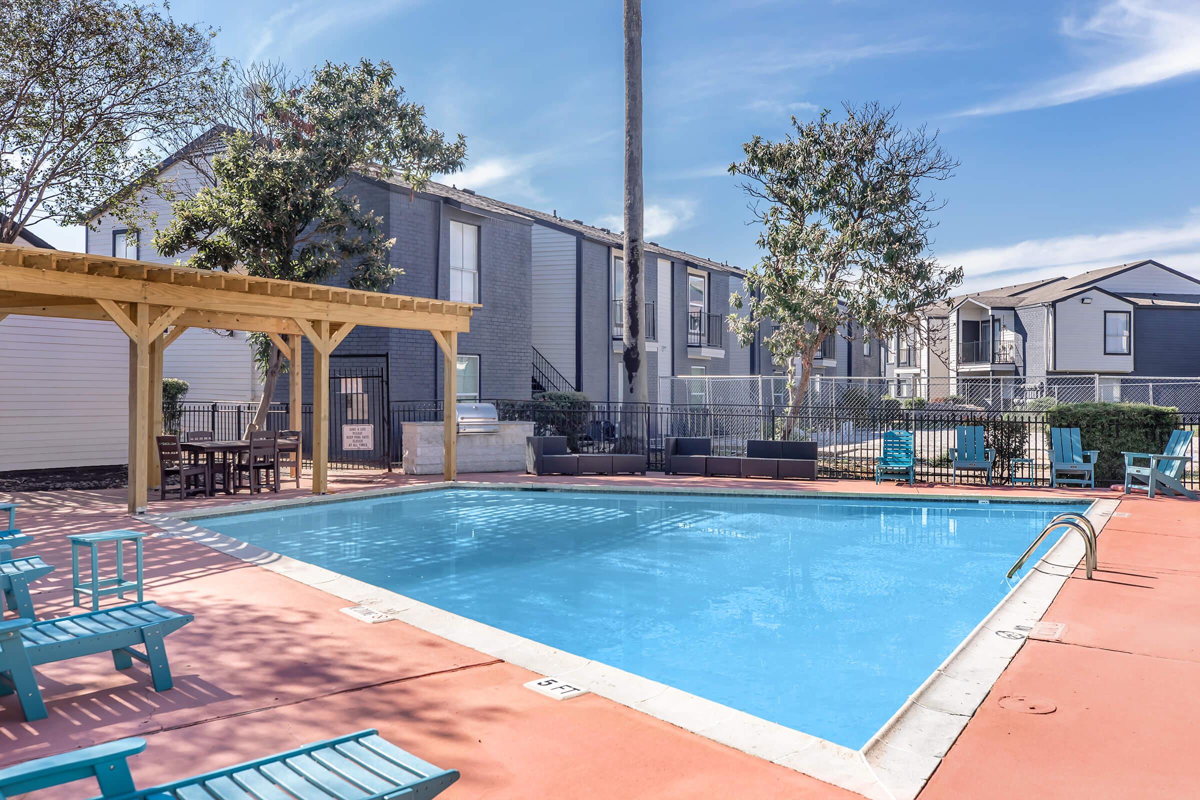 A swimming pool surrounded by a deck with teal chairs and tables. There are shaded areas with wooden structures, and apartment buildings in the background. The pool area is well-lit with blue water reflecting the sky, creating a relaxing outdoor environment.