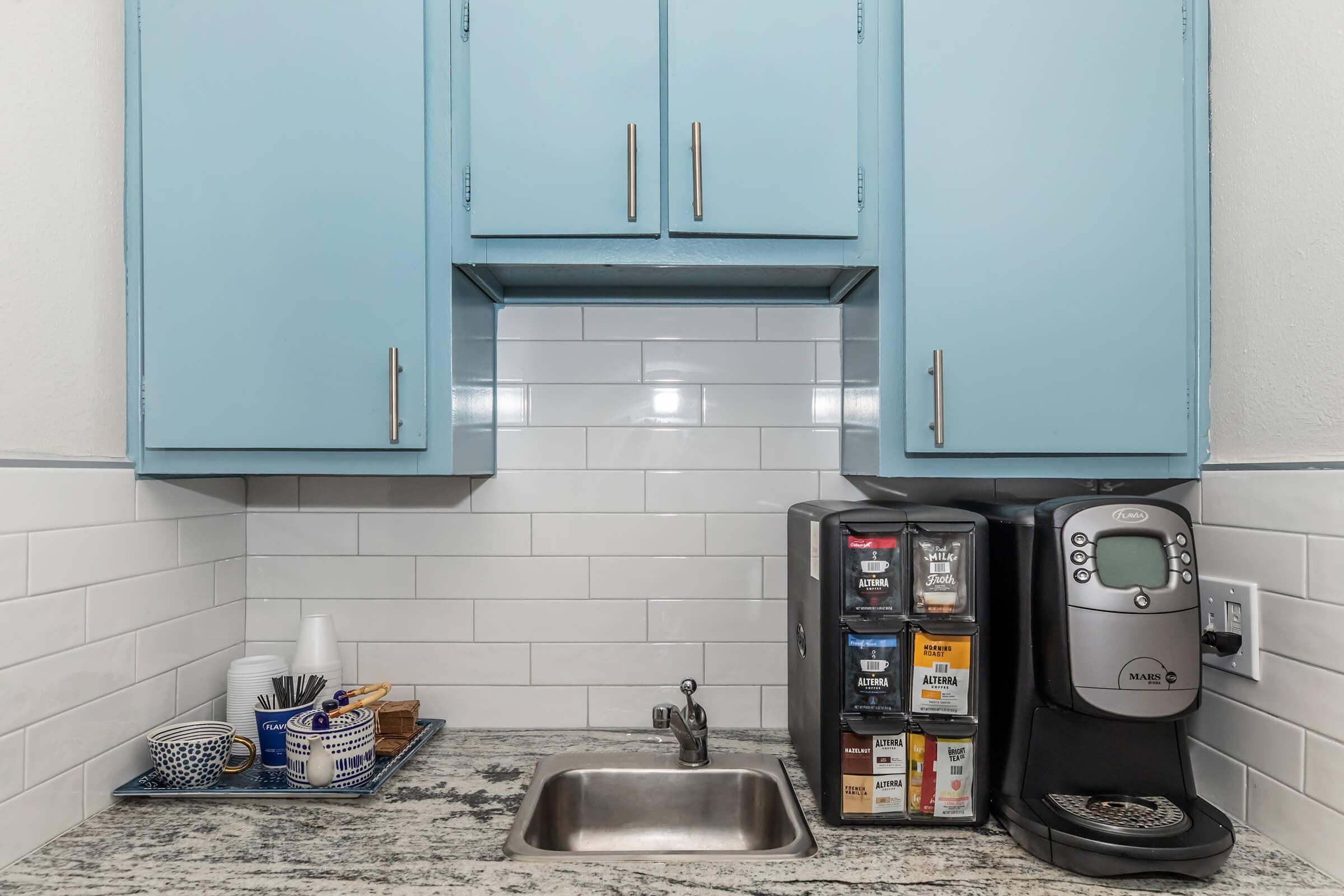 A small kitchen area featuring light blue cabinets, a stainless steel sink, and a coffee maker situated beside a stack of coffee pods. The countertop is made of granite with various kitchen items, including cups and a plate, arranged neatly. The wall behind is tiled with white subway tiles.