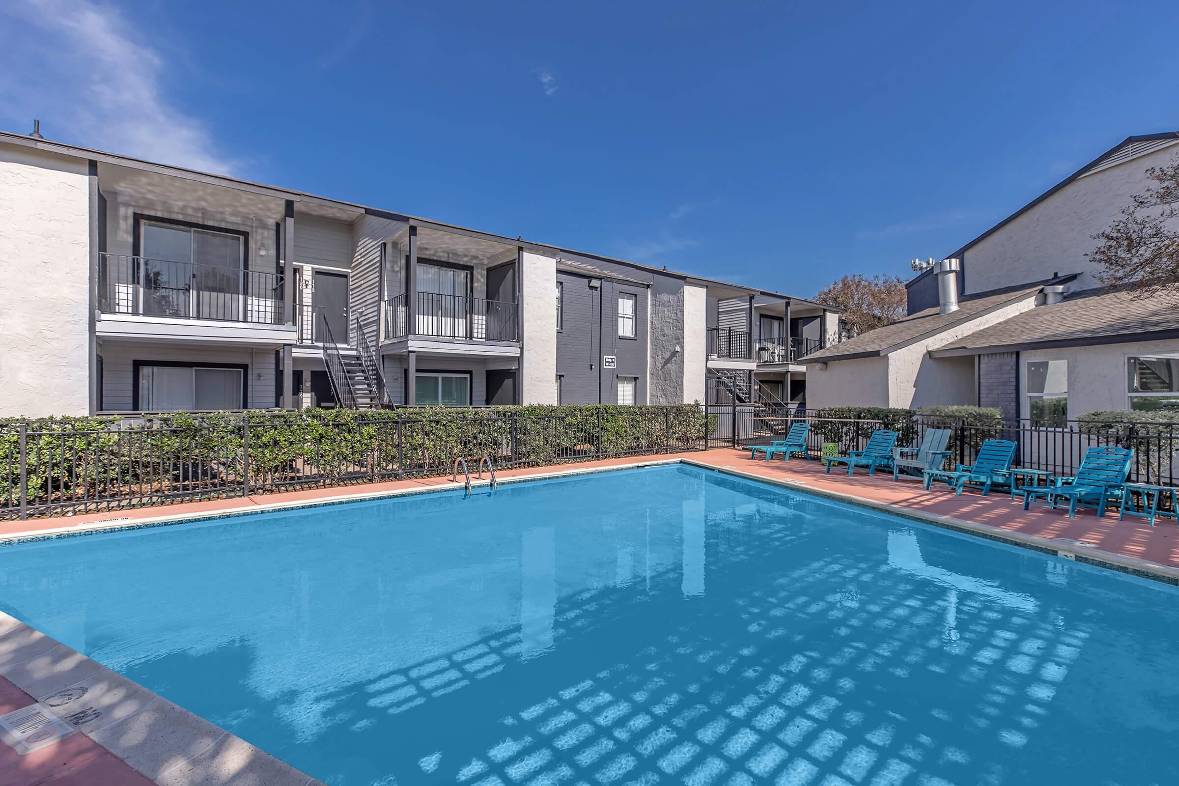 A clear blue swimming pool surrounded by a fenced area, with several lounge chairs on one side. In the background, there are two-story apartment buildings with balconies. The sky is bright blue with a few clouds, creating a pleasant atmosphere.