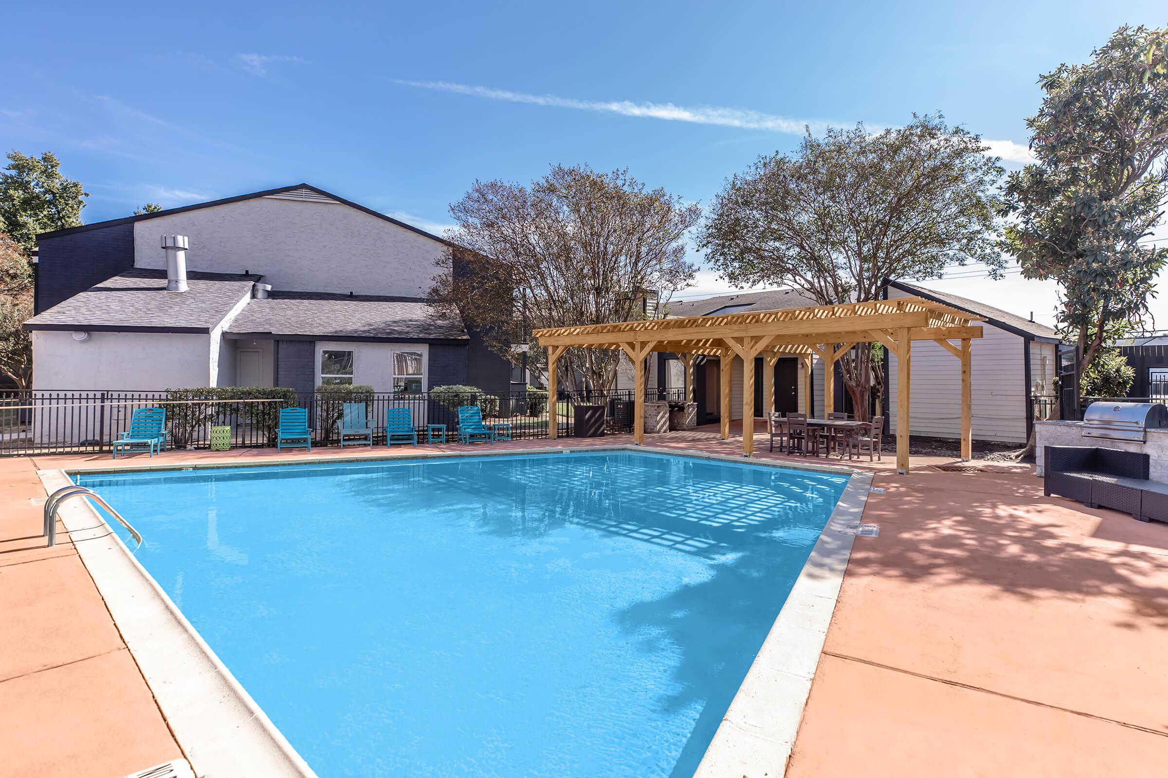 A clear blue swimming pool surrounded by a landscaped area. There are lounge chairs placed around the pool, a shaded pergola with seating, and a barbecue area nearby. The background features a modern building and trees under a bright blue sky.