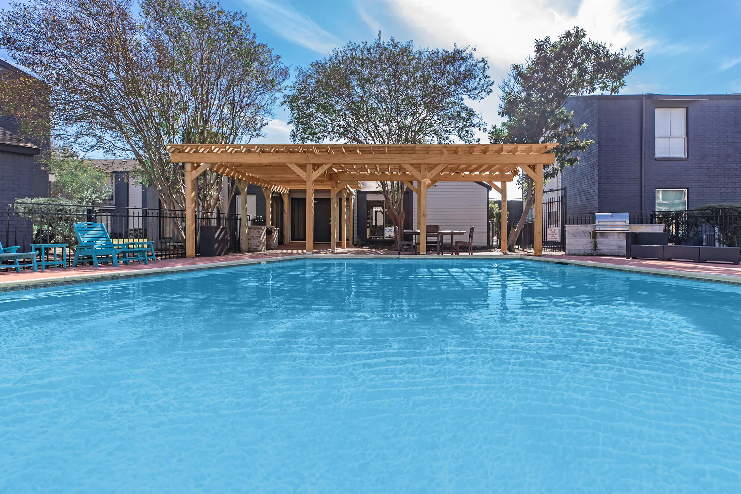 A clear swimming pool with a wooden pergola nearby, surrounded by trees and residential buildings. Blue lounge chairs are positioned around the pool area, creating a relaxing outdoor atmosphere on a sunny day.