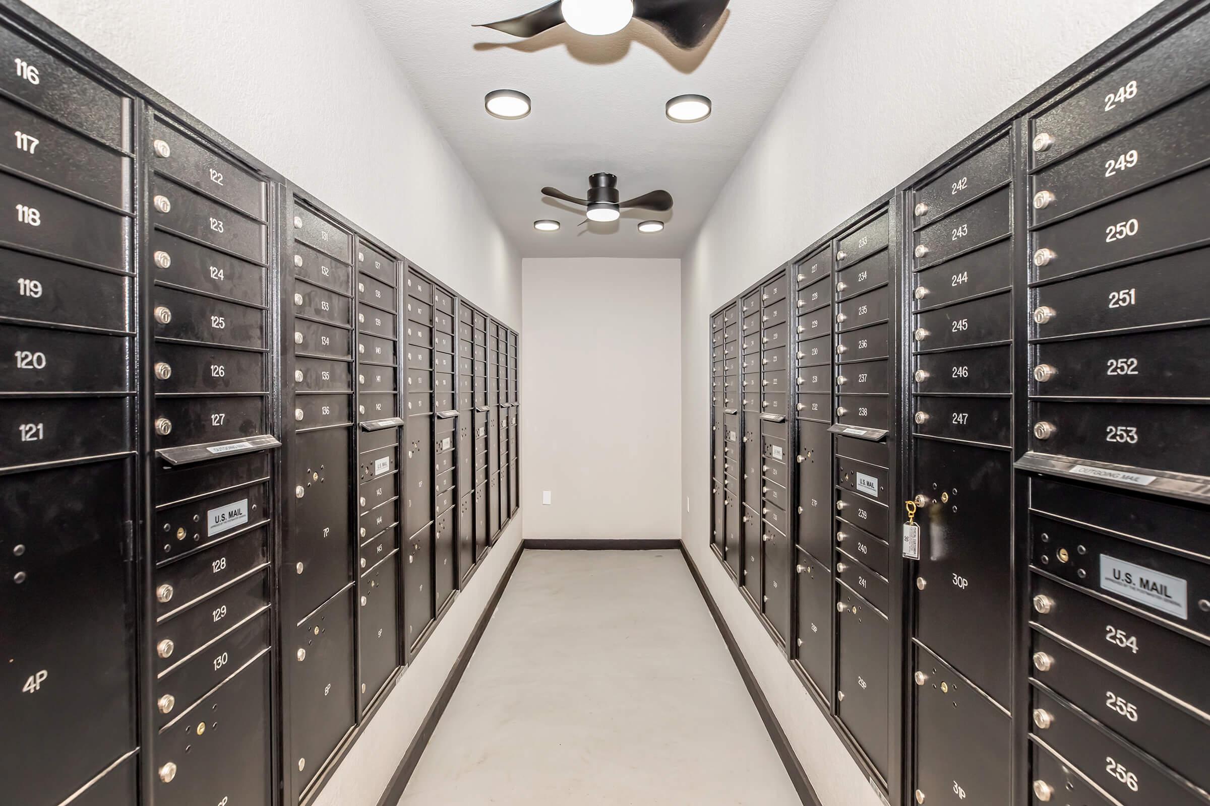 A corridor lined with black mailboxes on both sides, featuring a ceiling with modern lights. The area is well-lit and has a clean, minimalistic design, suggesting a residential or commercial space for securely storing mail.