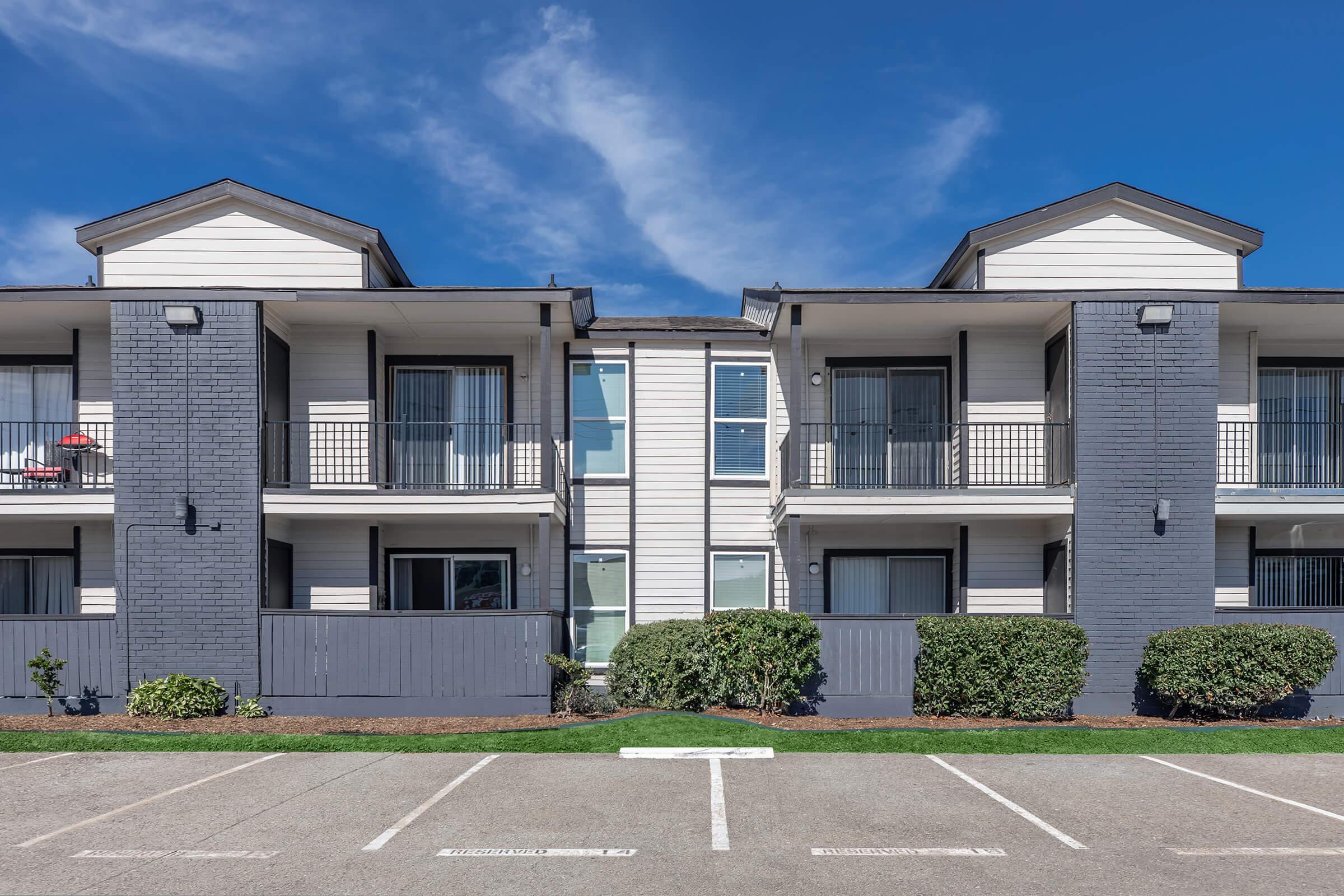 Two-story apartment building with gray and white exteriors. Each unit features a balcony with black railings. Green landscaping lines the front, and a paved parking lot is visible in the foreground. The sky is bright blue with a few clouds.