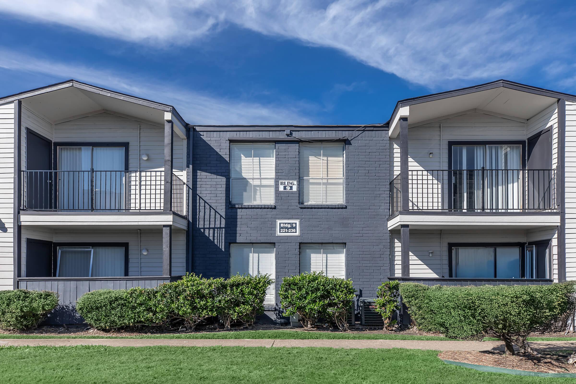 Two-story apartment building with a dark gray center section and light-colored side wings. Each side has a balcony with white railings. The building is surrounded by well-maintained green shrubs and grass. The sky is clear with some clouds.