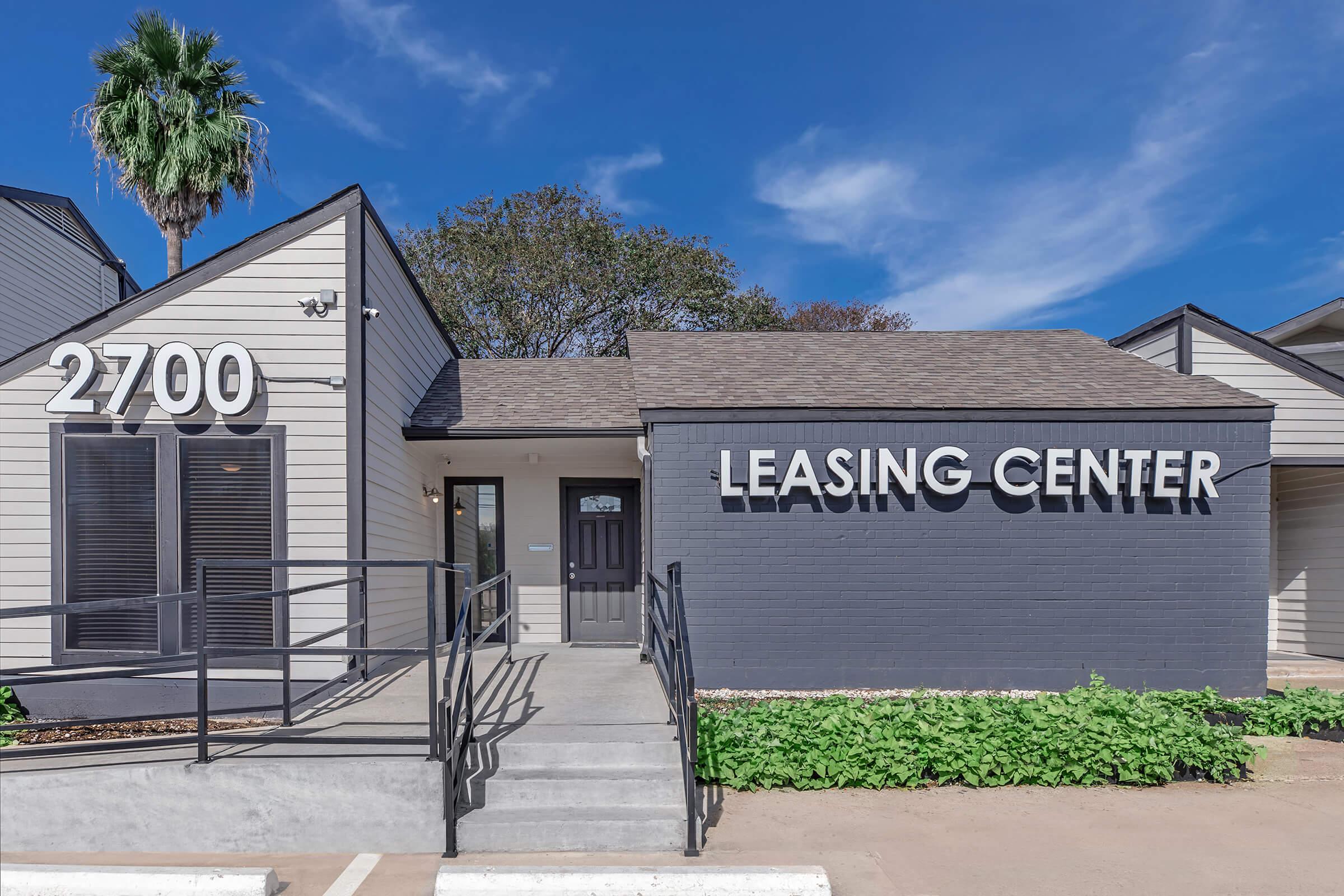 Leasing center of an apartment complex featuring a modern design. The building has a sloped roof, a large sign displaying "LEASING CENTER," and a concrete pathway leading to the entrance. Surrounding greenery and a palm tree provide a welcoming atmosphere. Clear blue sky in the background.