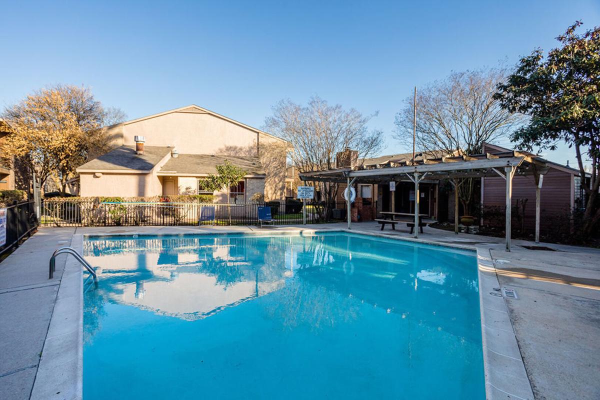 A clear blue swimming pool surrounded by a concrete deck, with a wooden pergola nearby. In the background, there are two residential buildings, and trees with sparse foliage. The sky is blue and clear, indicating a sunny day.